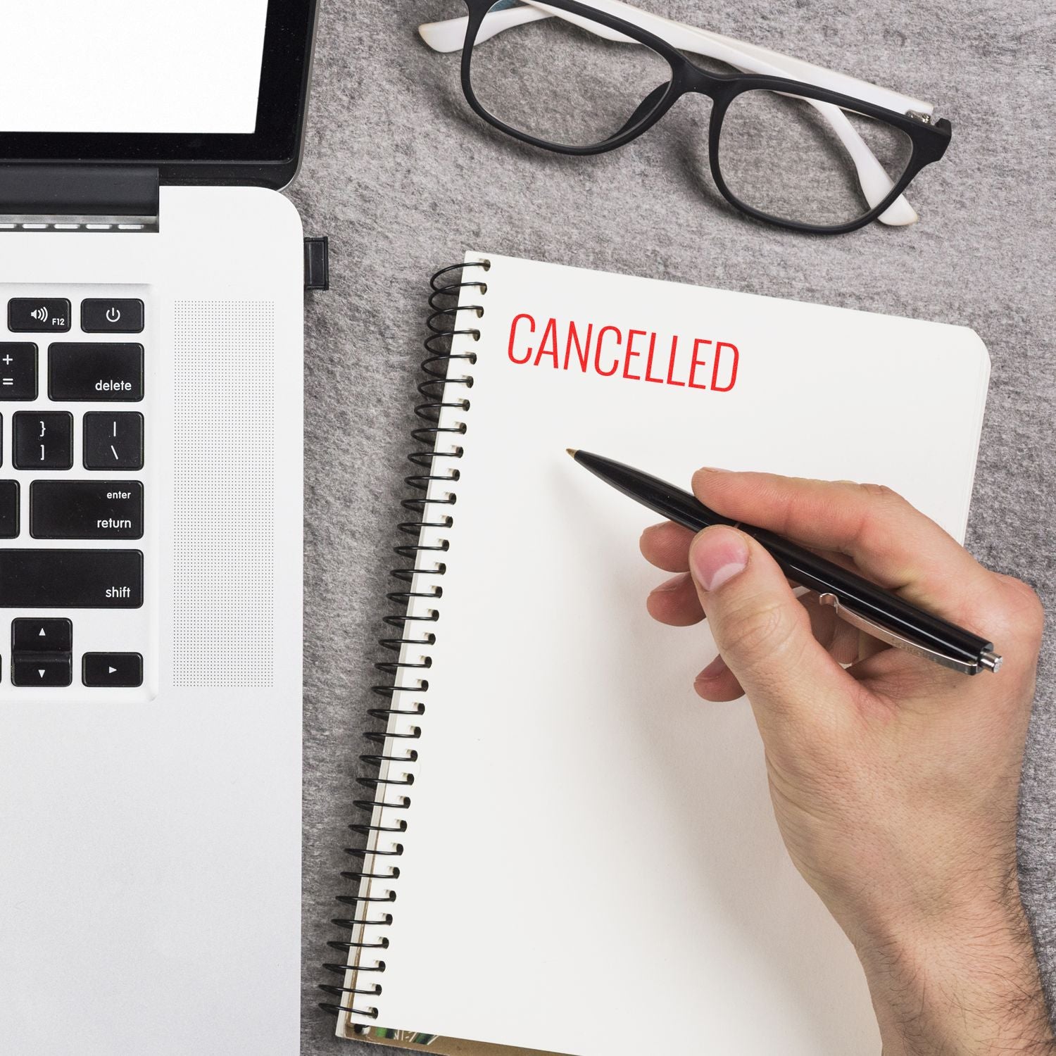 Hand holding pen near notebook with CANCELLED in narrow font, next to a laptop and glasses on a gray surface.