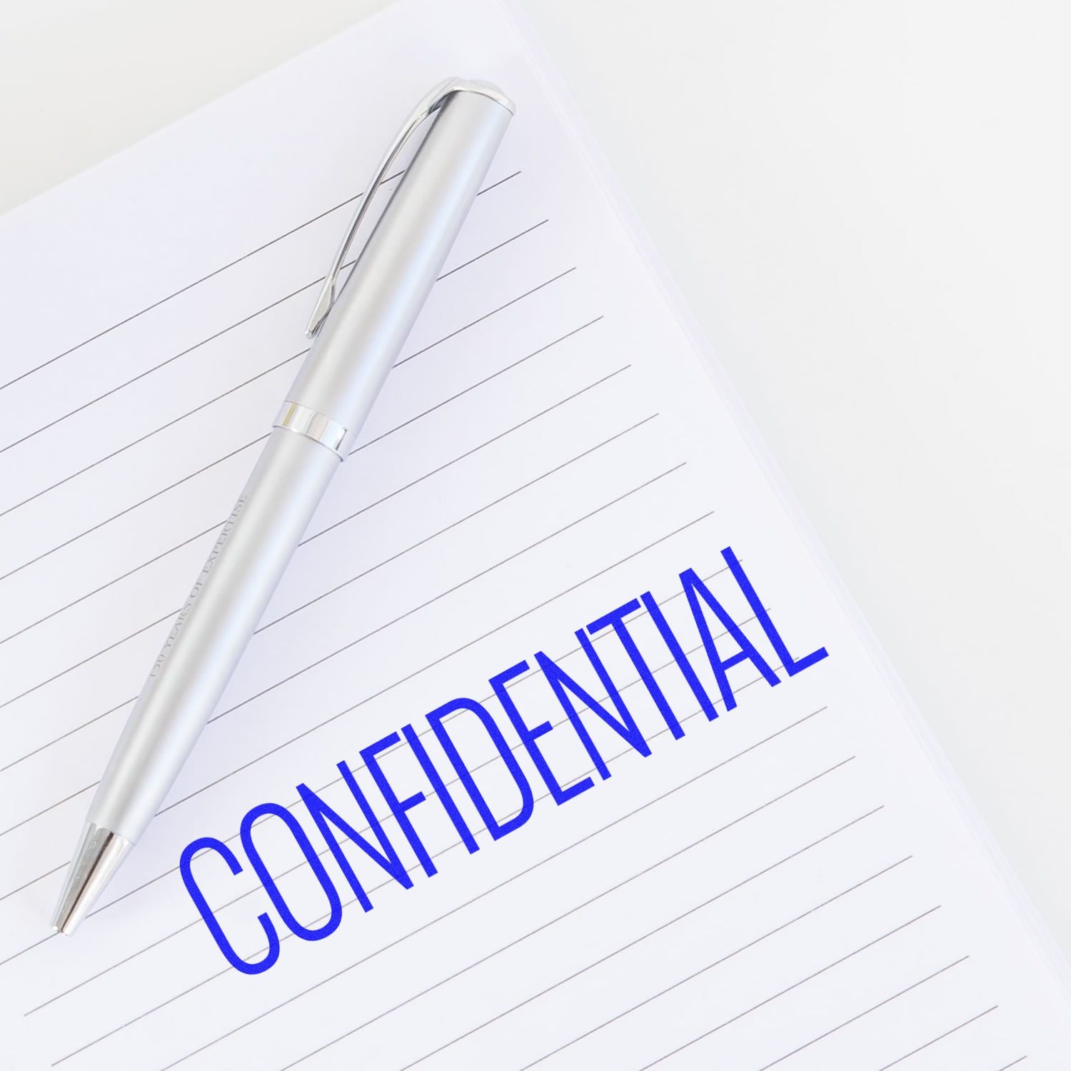 A silver pen lies on a lined notebook with the word "CONFIDENTIAL" stamped in blue using the Narrow Font Confidential Rubber Stamp.