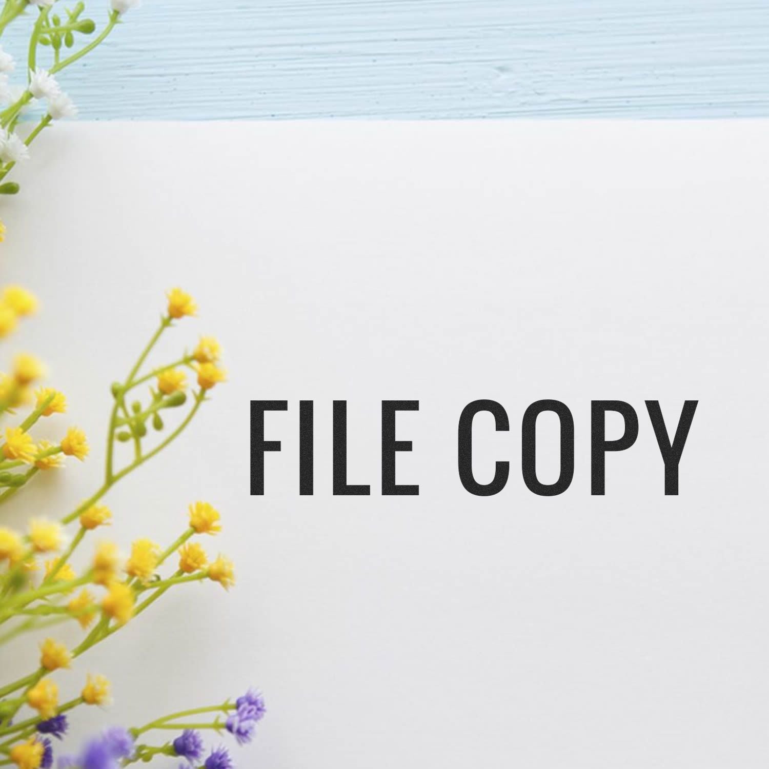 Large Narrow Font File Copy Rubber Stamp imprint on white paper, surrounded by yellow and purple flowers on a light blue background.