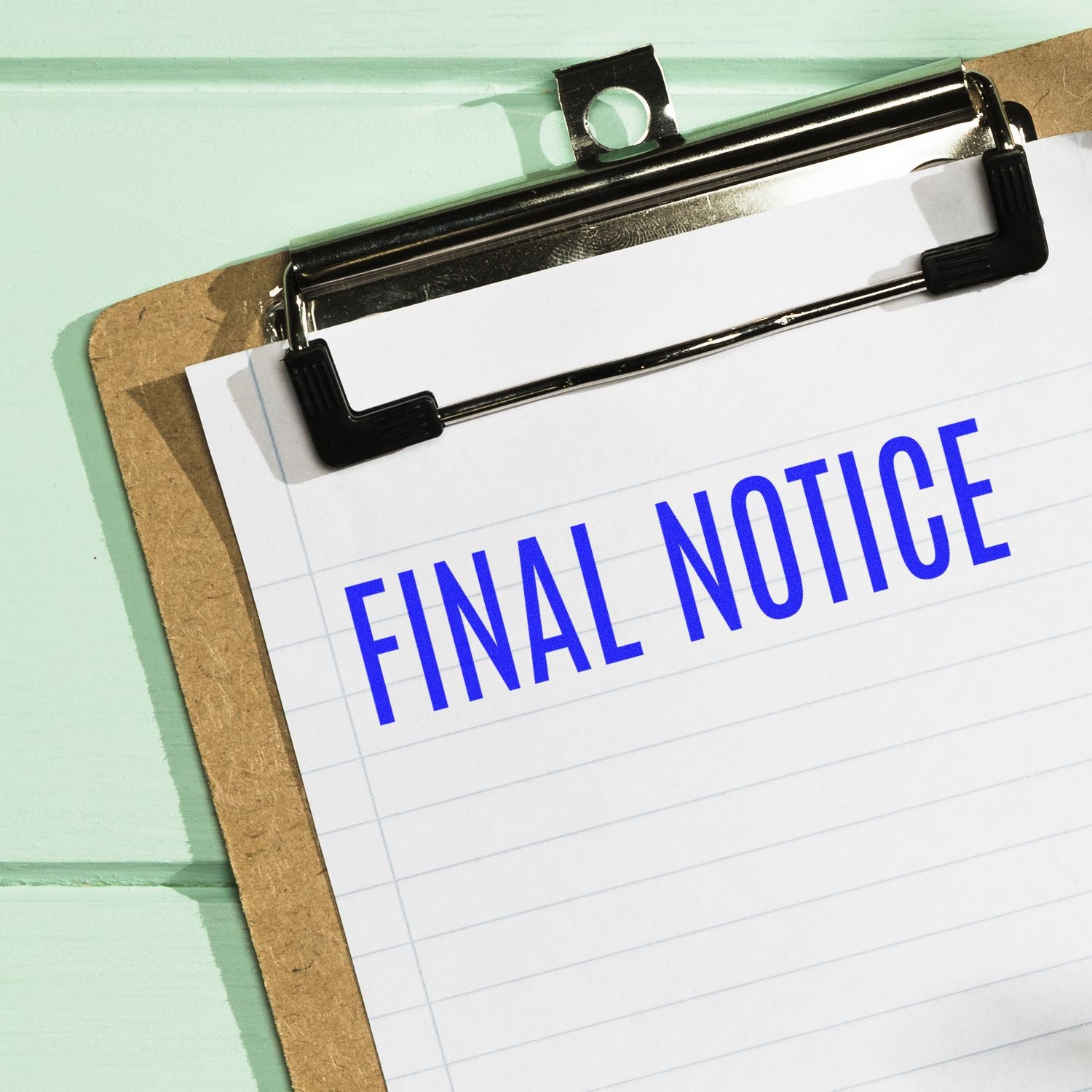 Clipboard with lined paper stamped "FINAL NOTICE" in blue using the Narrow Font Final Notice Rubber Stamp, placed on a mint green background.