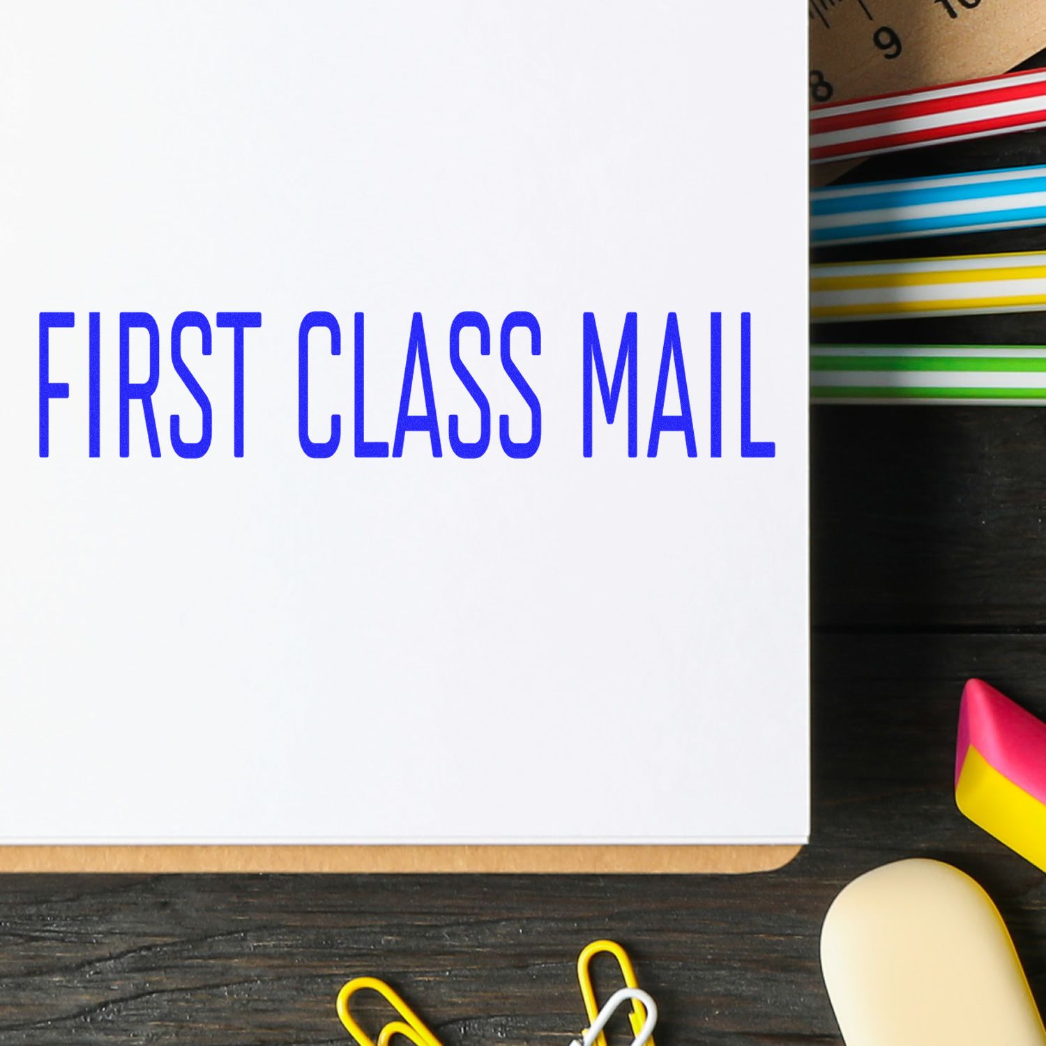 Narrow Font First Class Mail Rubber Stamp in blue ink on white paper, surrounded by colorful office supplies on a wooden desk.