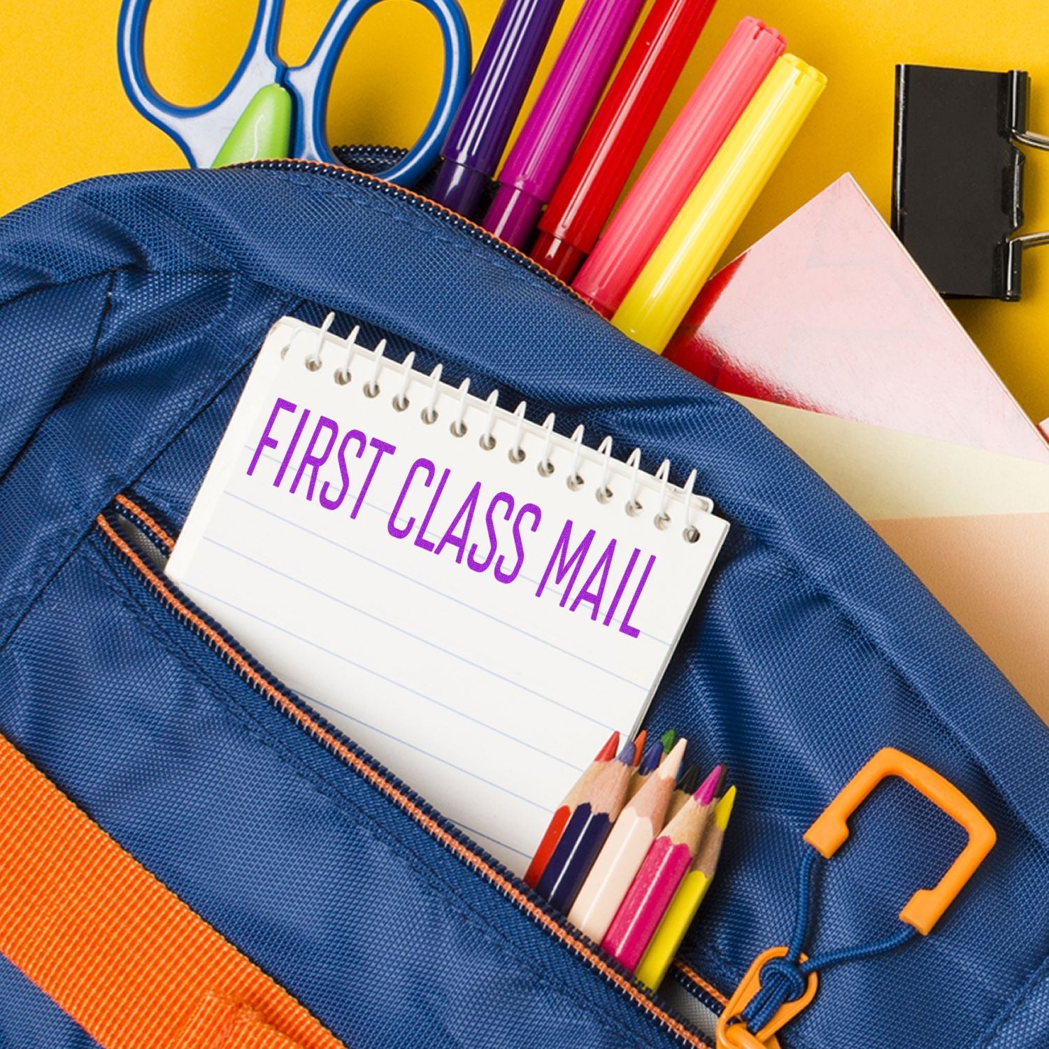 A notepad with First Class Mail in a narrow font rubber stamp is placed in the front pocket of a blue backpack with school supplies.