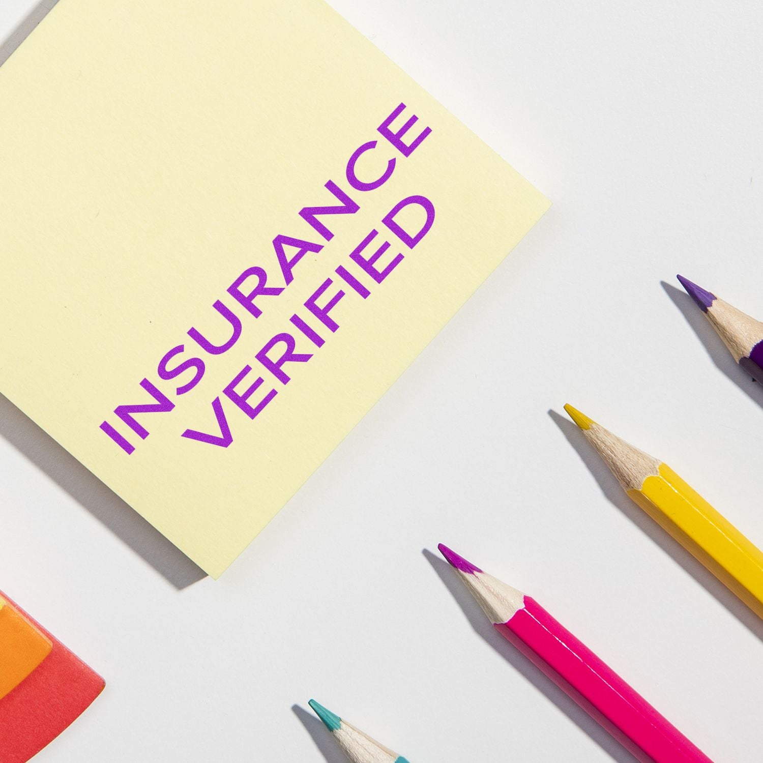 Yellow sticky note with Insurance Verified in narrow font, surrounded by colorful pencils and an eraser on a white background.