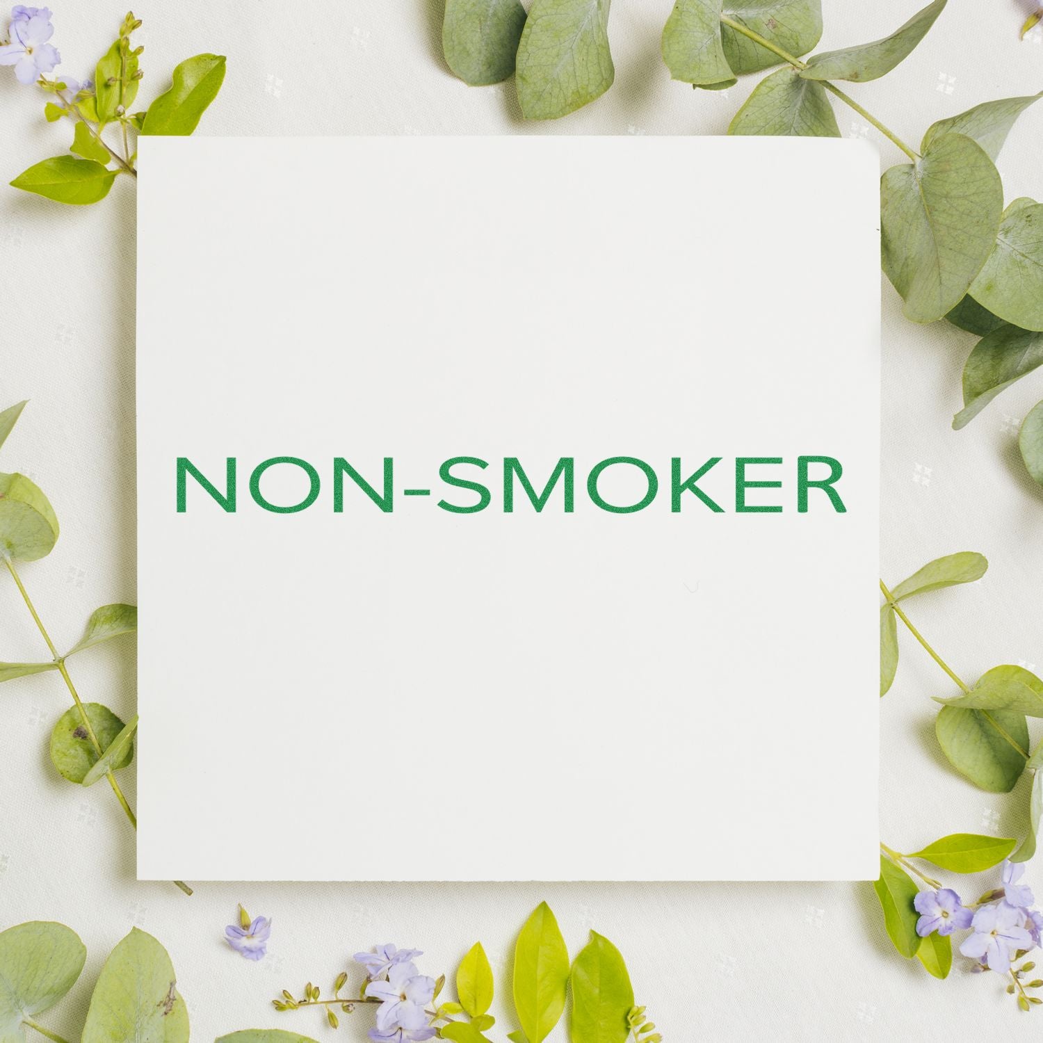 Narrow Font Non-Smoker Rubber Stamp on white paper surrounded by green leaves and small purple flowers.