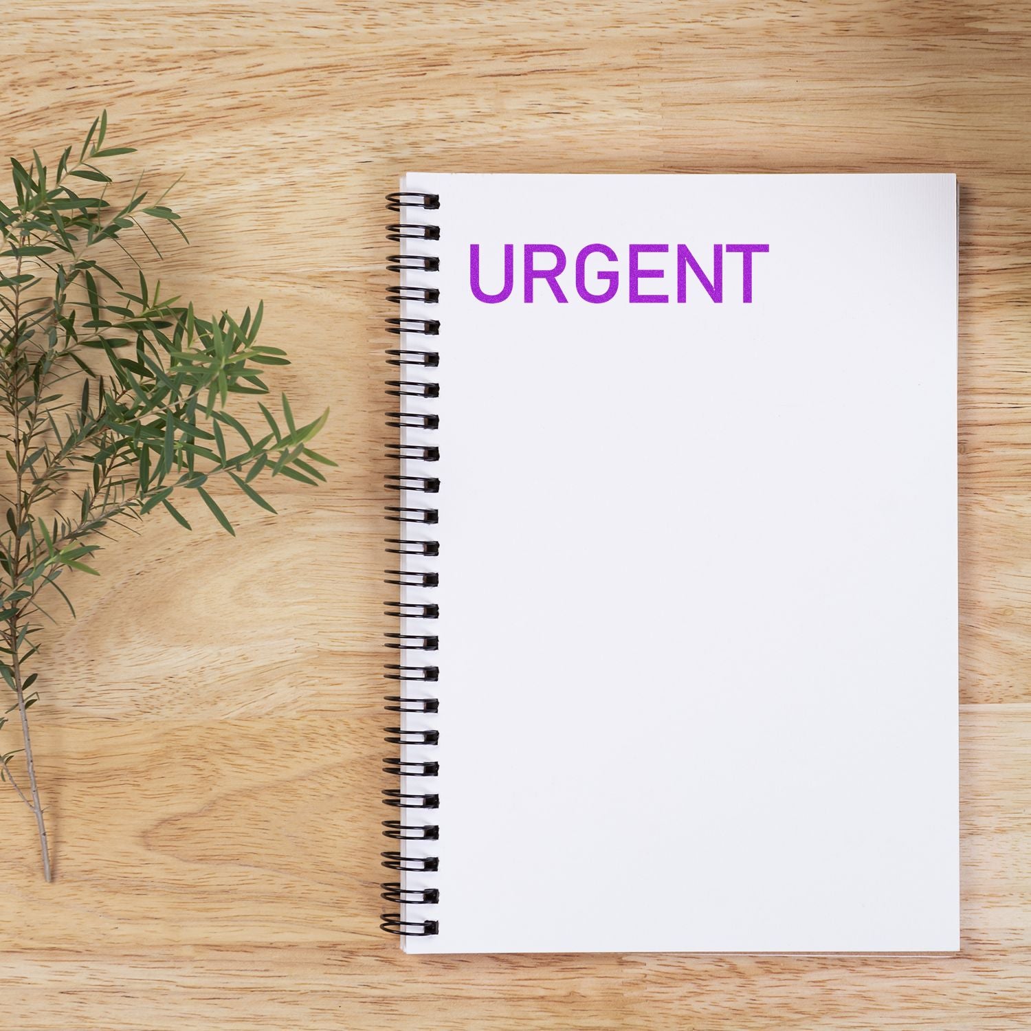 Narrow Font Urgent Rubber Stamp in purple ink on a white spiral notebook, placed on a wooden surface with a plant.