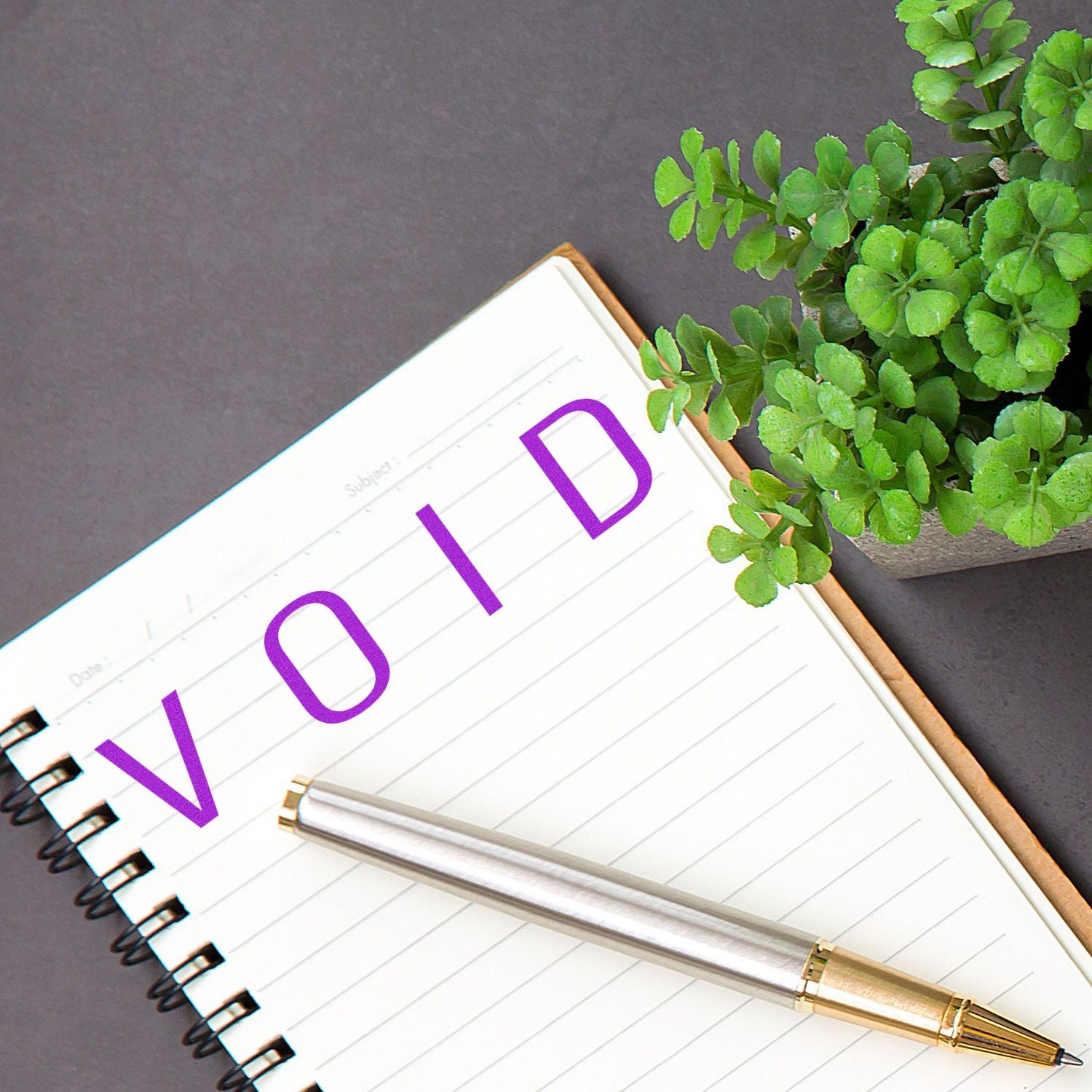 Notebook stamped with 'VOID' using a Slim Pre-Inked Narrow Void Stamp, next to a silver pen and a potted plant.