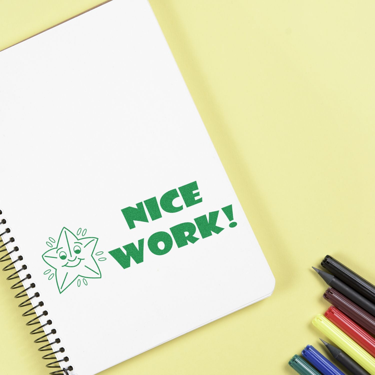 Nice Work Rubber Stamp on a spiral notebook with colorful pens on a yellow background.