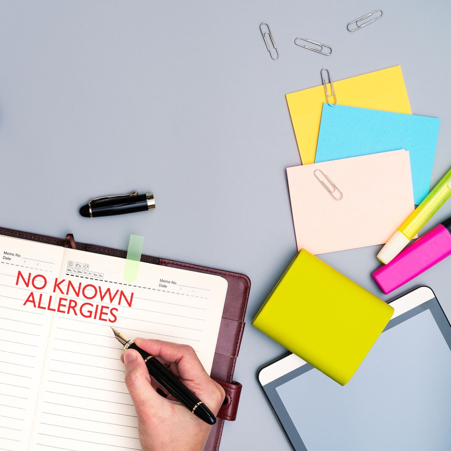 A hand using the Large No Known Allergies Rubber Stamp on a notebook, surrounded by colorful stationery, a tablet, and a pen.