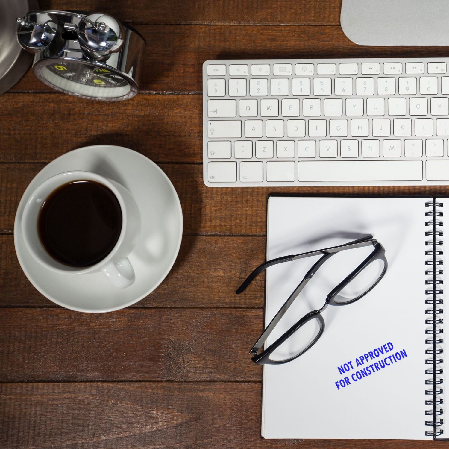 A desk with a keyboard, coffee, glasses, and a notebook stamped with Not Approved For Construction using a Slim Pre-Inked Stamp.