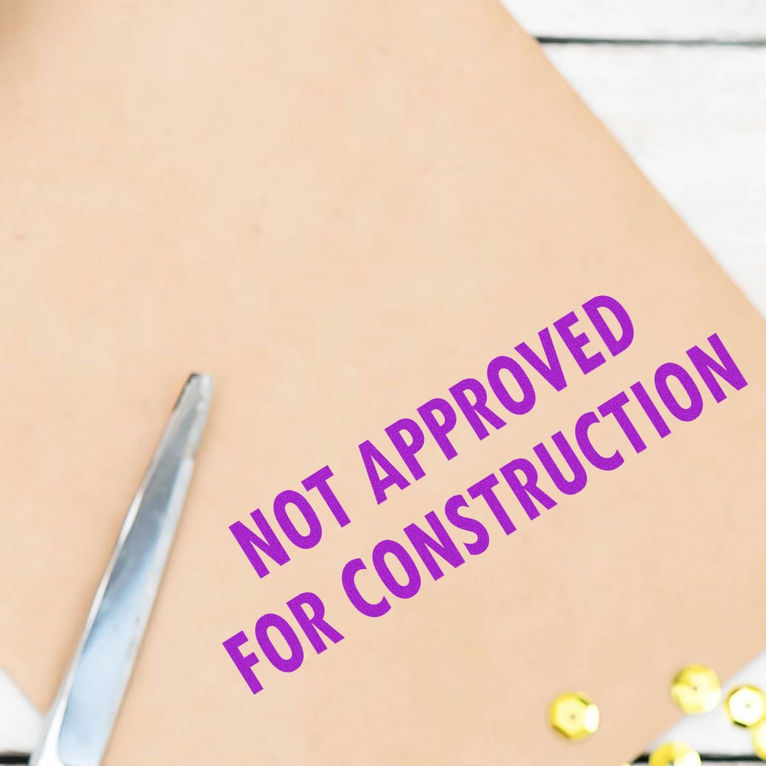 Not Approved For Construction rubber stamp in purple ink on a beige paper with a pair of scissors and gold buttons nearby.