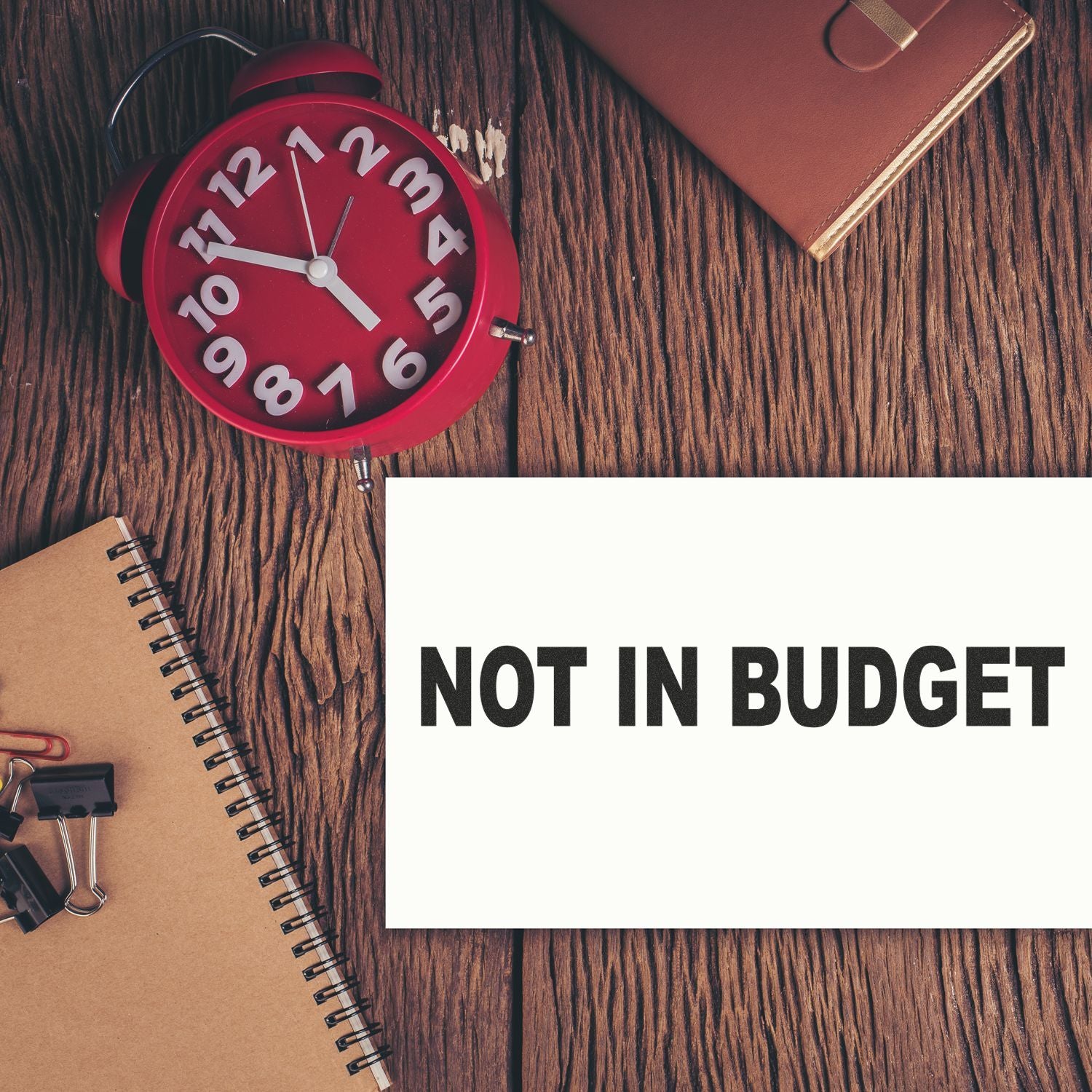 Large Self Inking Not In Budget Stamp on a wooden desk with a red clock, notebook, and binder clips.