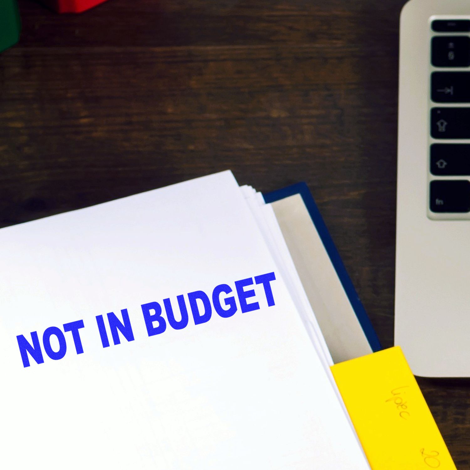 Large Self Inking Not In Budget Stamp imprint on a document beside a laptop and sticky notes on a wooden desk.