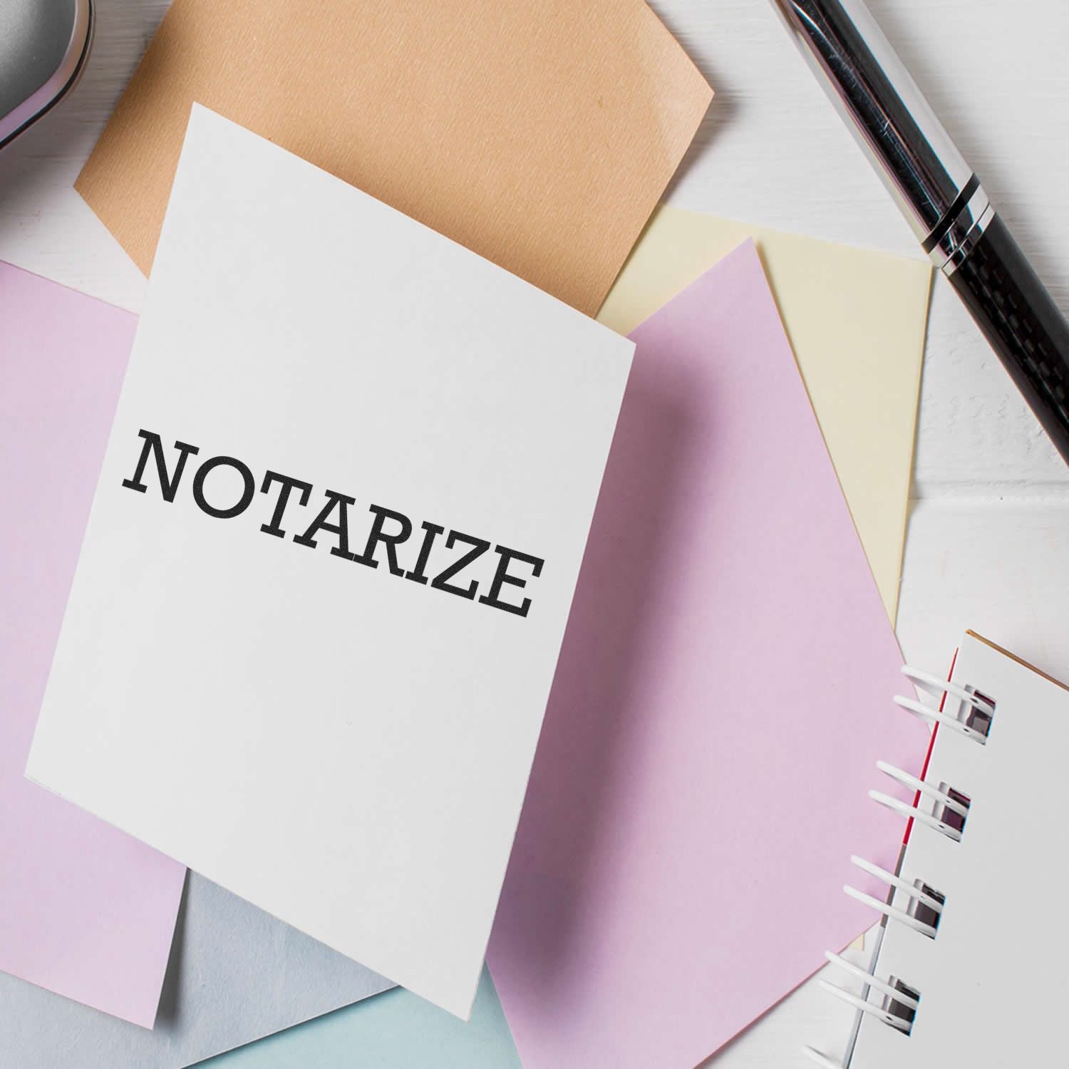 Large Notarize Rubber Stamp in use on a white paper, surrounded by colorful papers, a pen, and a notebook on a white desk.