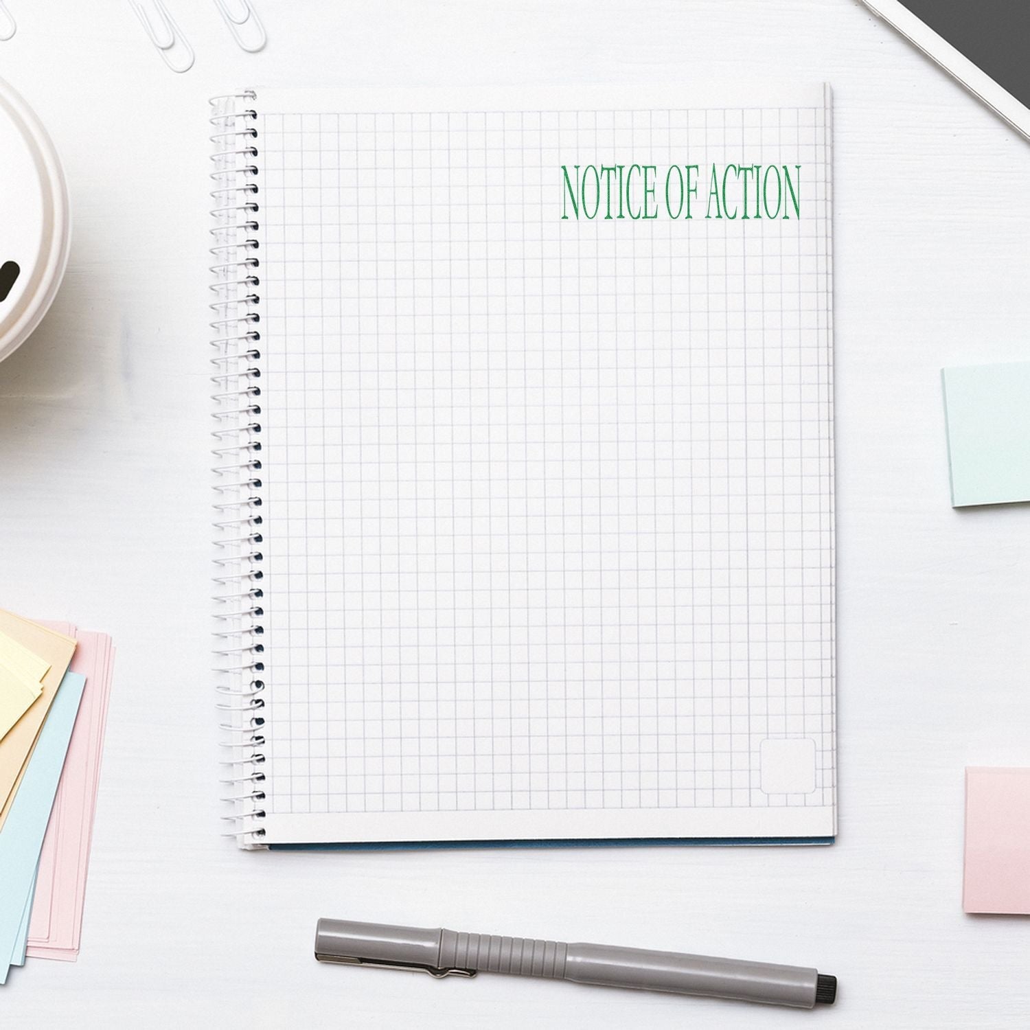 A notebook with a grid pattern stamped with NOTICE OF ACTION in green using the Self Inking Notice Of Action Stamp, surrounded by office supplies.