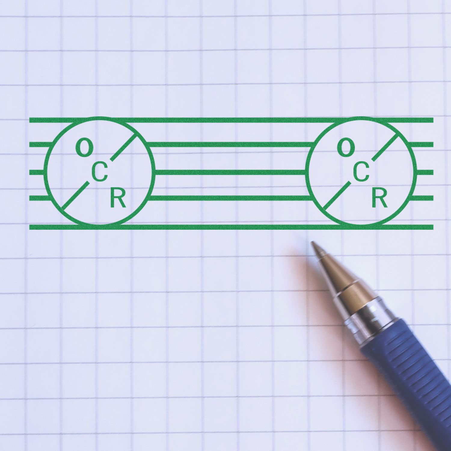 A blue pen next to a green stamped design on graph paper using the OCR Rubber Stamp, featuring circles with "OCR" text and horizontal lines.