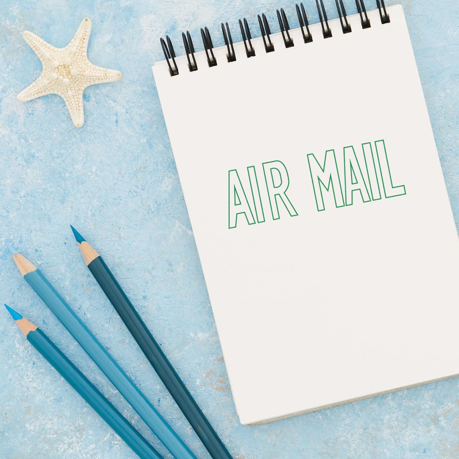 Notebook stamped with AIR MAIL using the Large Outline Air Mail Rubber Stamp, placed on a blue surface with pencils and a starfish.