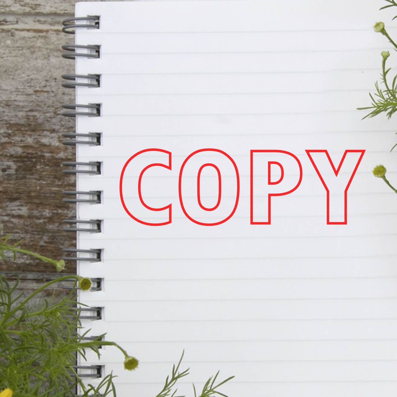 Large Self Inking Outline Copy Stamp in red ink on a spiral notebook with a wooden background and green plants.