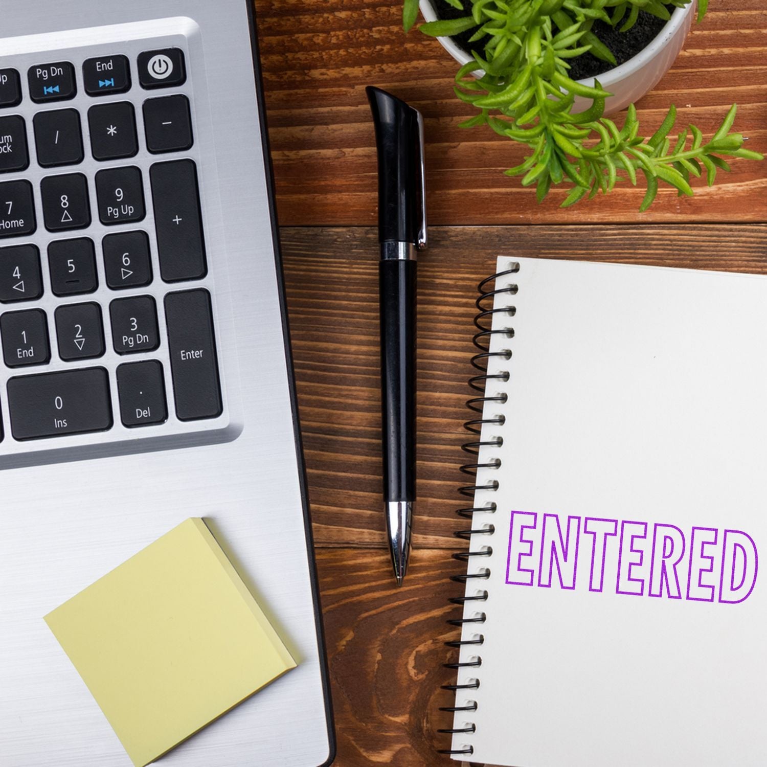 A desk with a laptop, sticky notes, pen, plant, and a notebook stamped with ENTERED using the Large Entered With Outline Rubber Stamp.