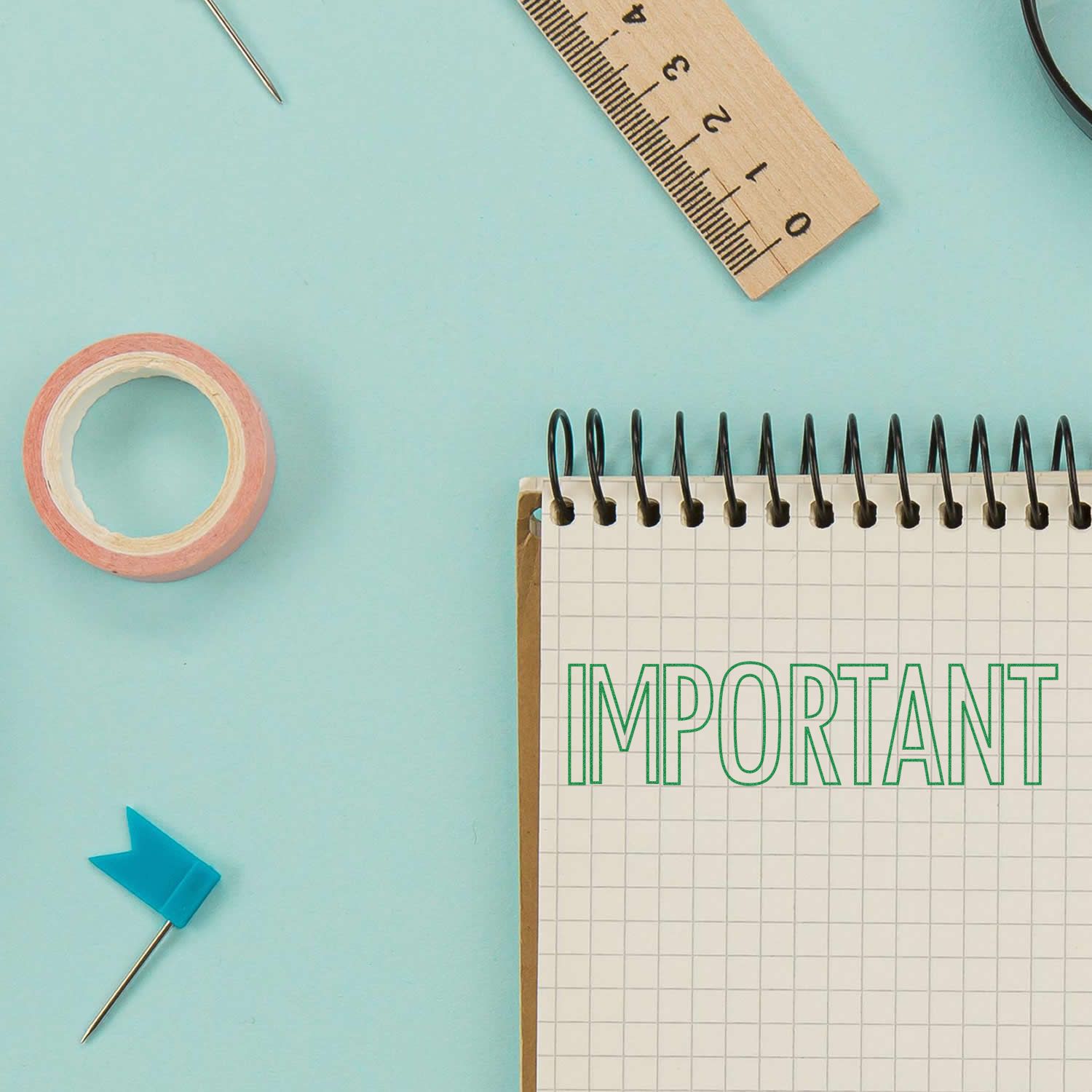 A Self Inking Outline Important Stamp is used on a spiral notebook. Nearby are a ruler, a roll of tape, a pin, and a needle on a blue surface.