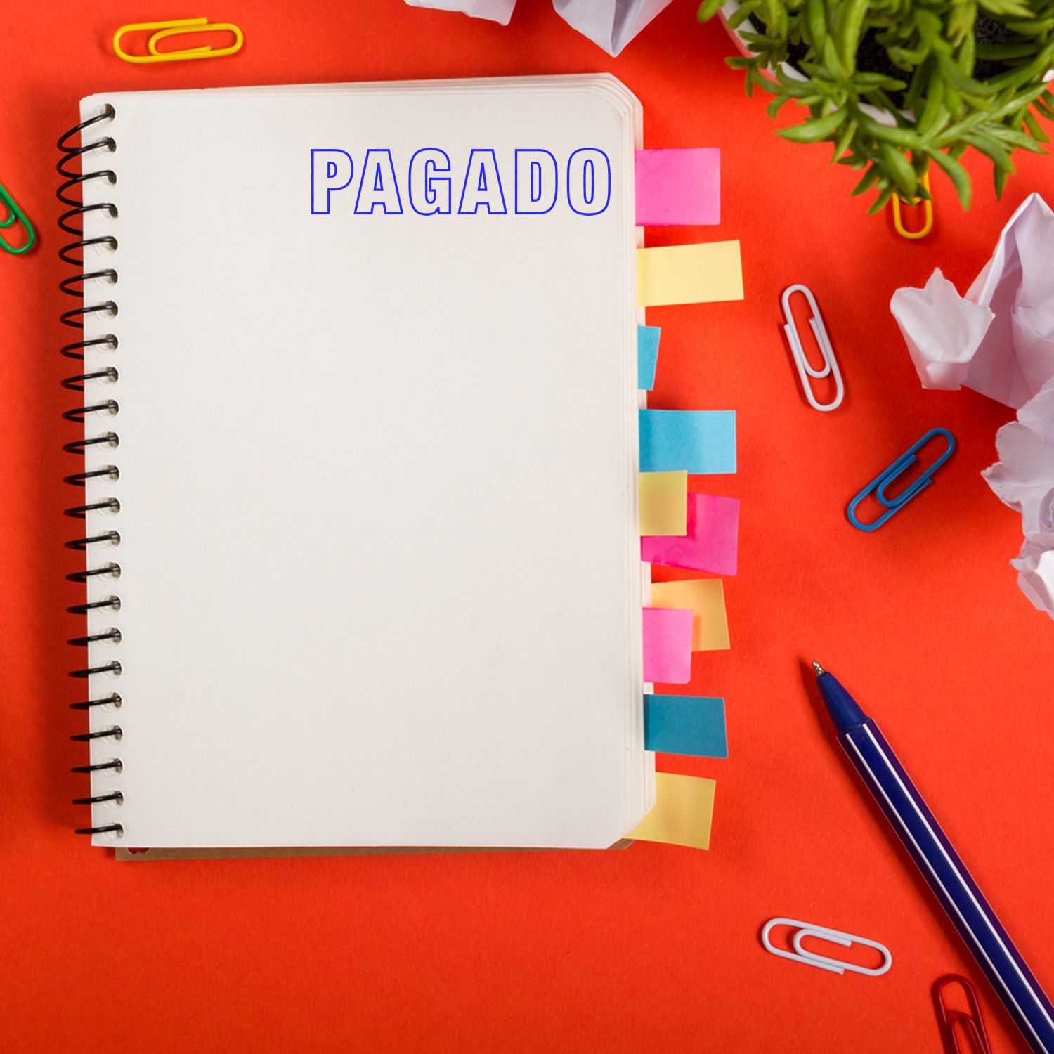 Self Inking Outline Pagado Stamp marking PAGADO on a notebook with colorful tabs, surrounded by paper clips, a pen, and a plant on a red background.