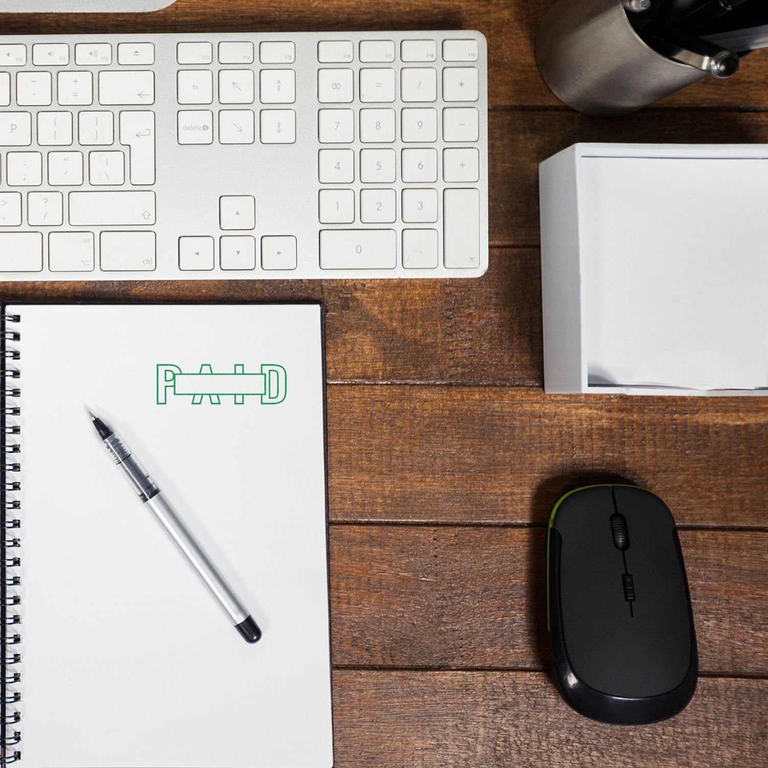 Notebook stamped with PAID using Outline Paid with Box Rubber Stamp, placed on a wooden desk with a keyboard, mouse, and pen.