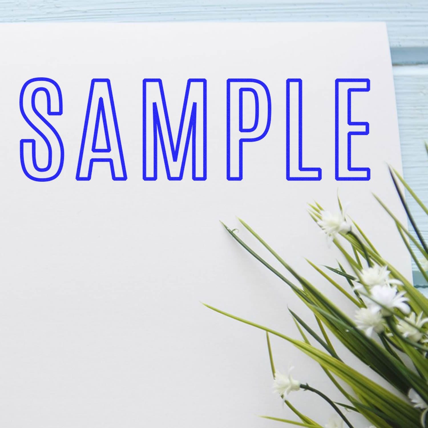 Large Self Inking Outline Sample Stamp imprinting the word 'SAMPLE' in blue on white paper, with green and white flowers beside it.