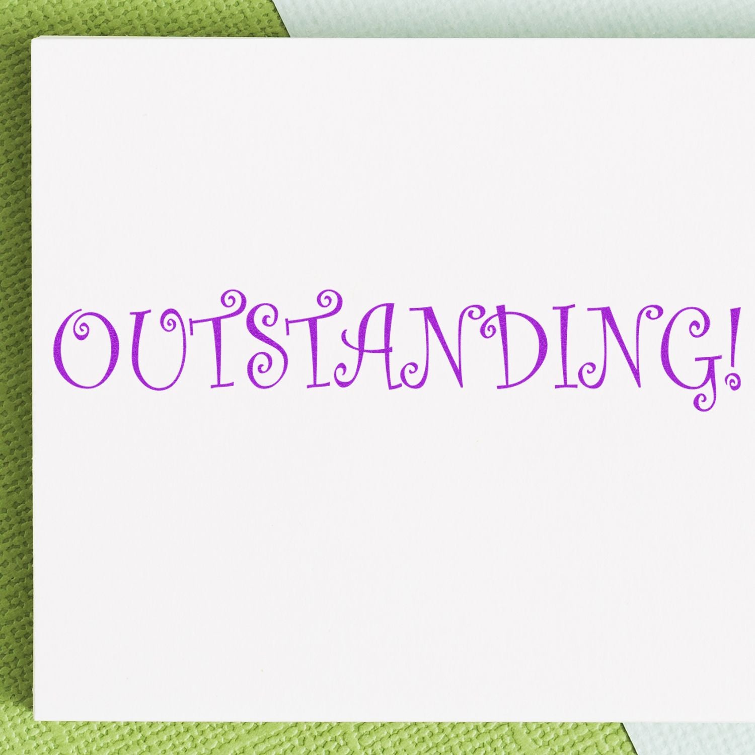 A white card stamped with OUTSTANDING! in purple using the Large Self Inking Outstanding Stamp, placed on a green textured surface.