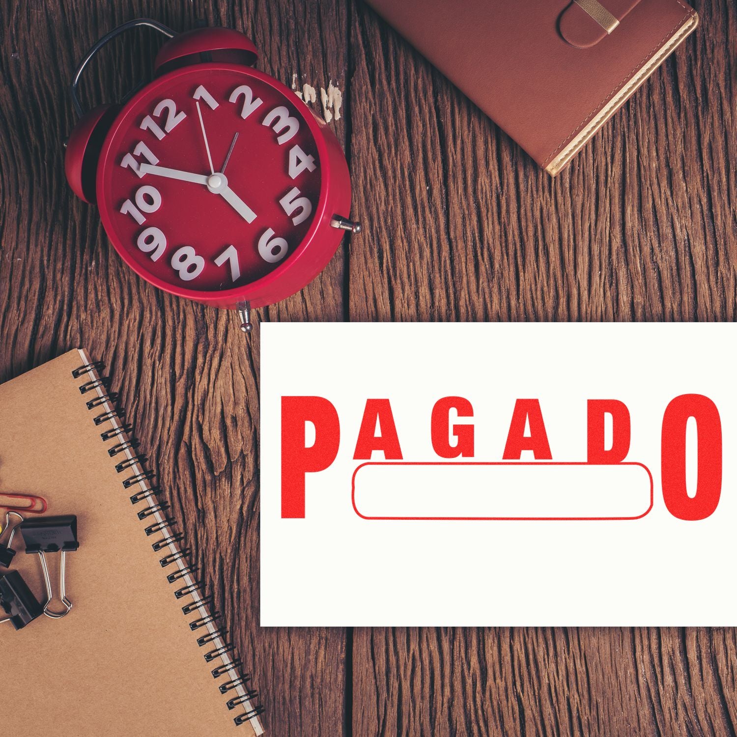 Pagado with Box Rubber Stamp on paper, surrounded by a red clock, notebook, and office supplies on a wooden desk.