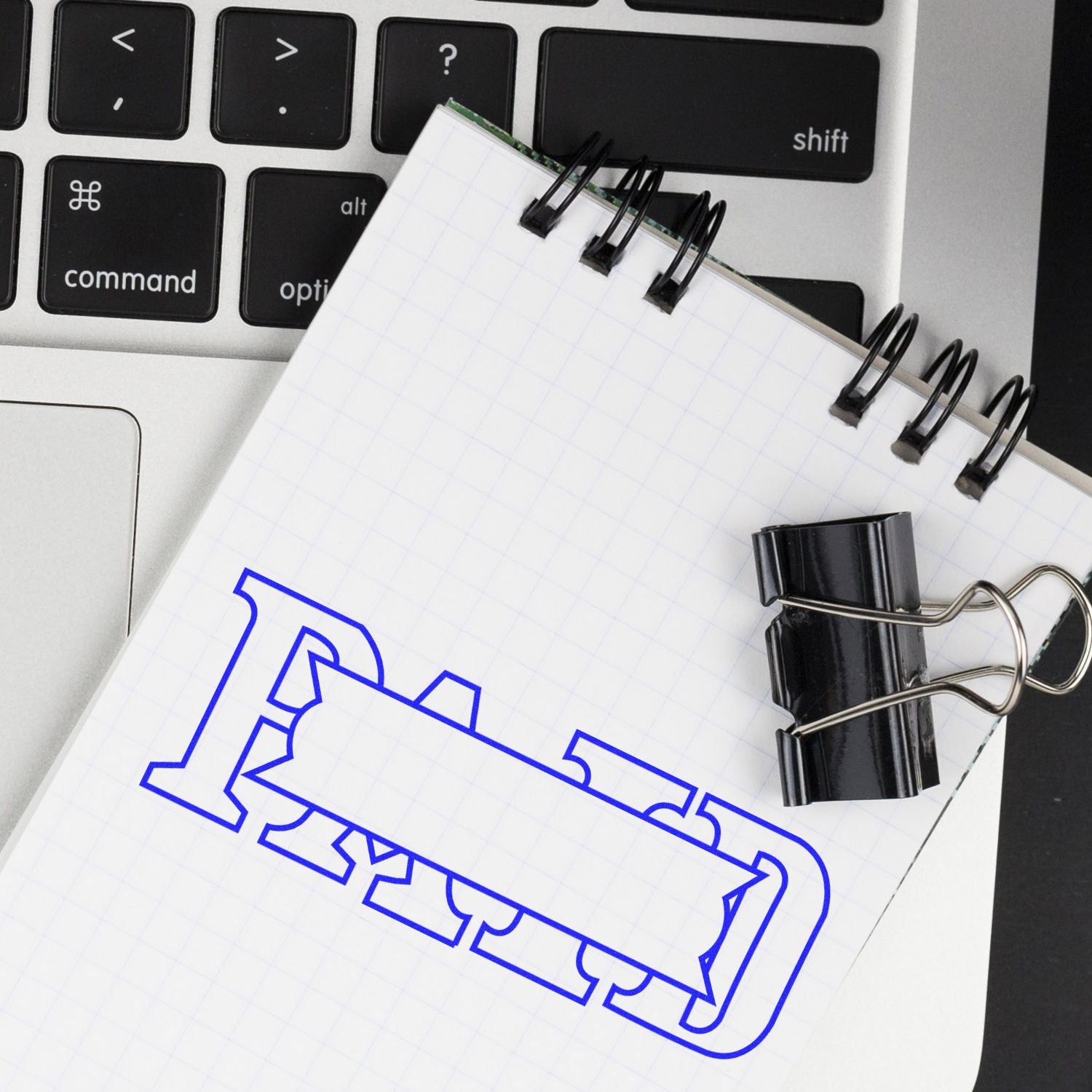 Notebook with 'Paid with Box Rubber Stamp' in blue ink on top of a laptop keyboard, held by a black binder clip.