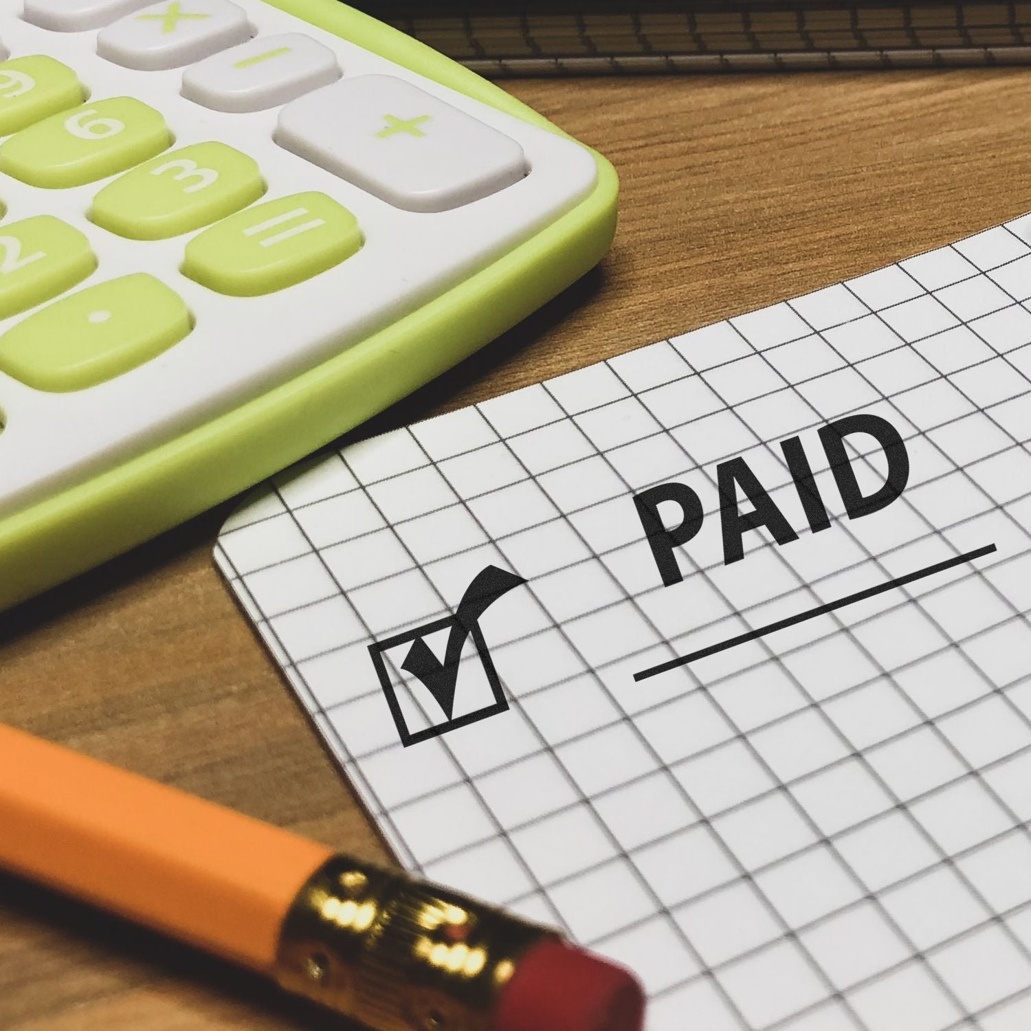 Large Paid with Checkmark Rubber Stamp used on graph paper next to a pencil and calculator on a wooden desk.