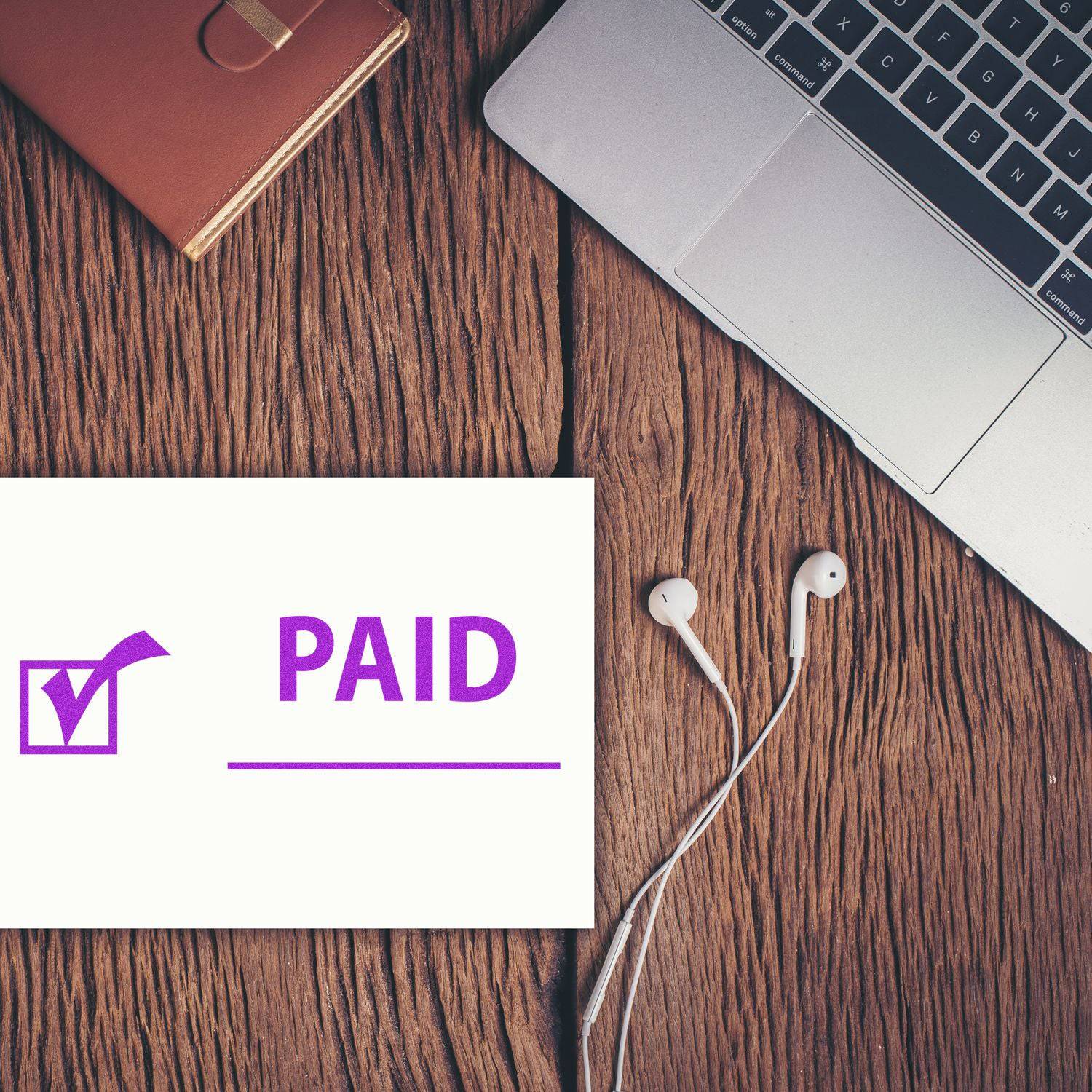 Large Paid with Checkmark Rubber Stamp in purple ink on paper, placed on a wooden desk next to a laptop, notebook, and earphones.