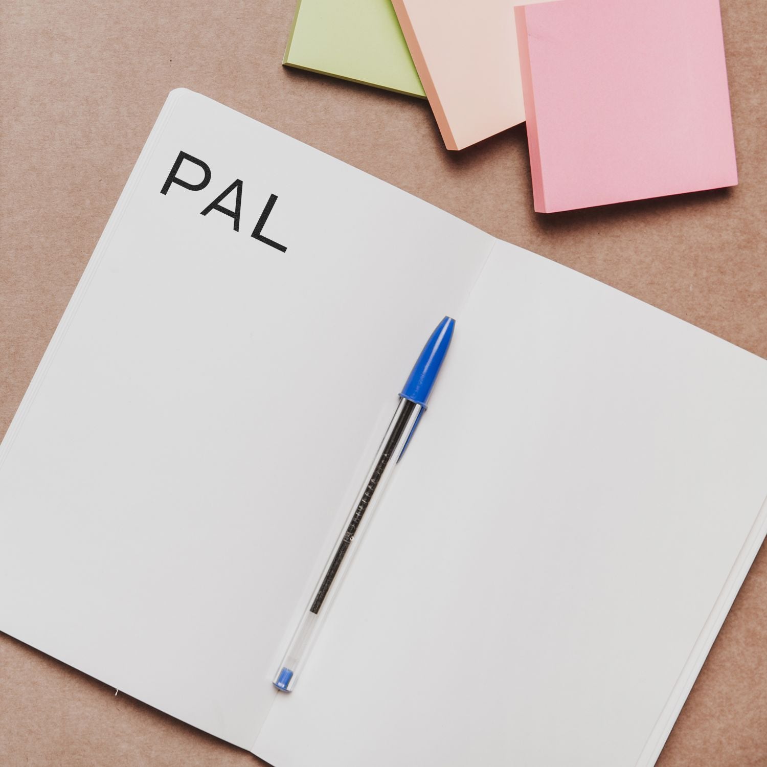 Open notebook stamped with PAL using Large Self Inking PAL Stamp, alongside a blue pen and colorful sticky notes on a brown surface.