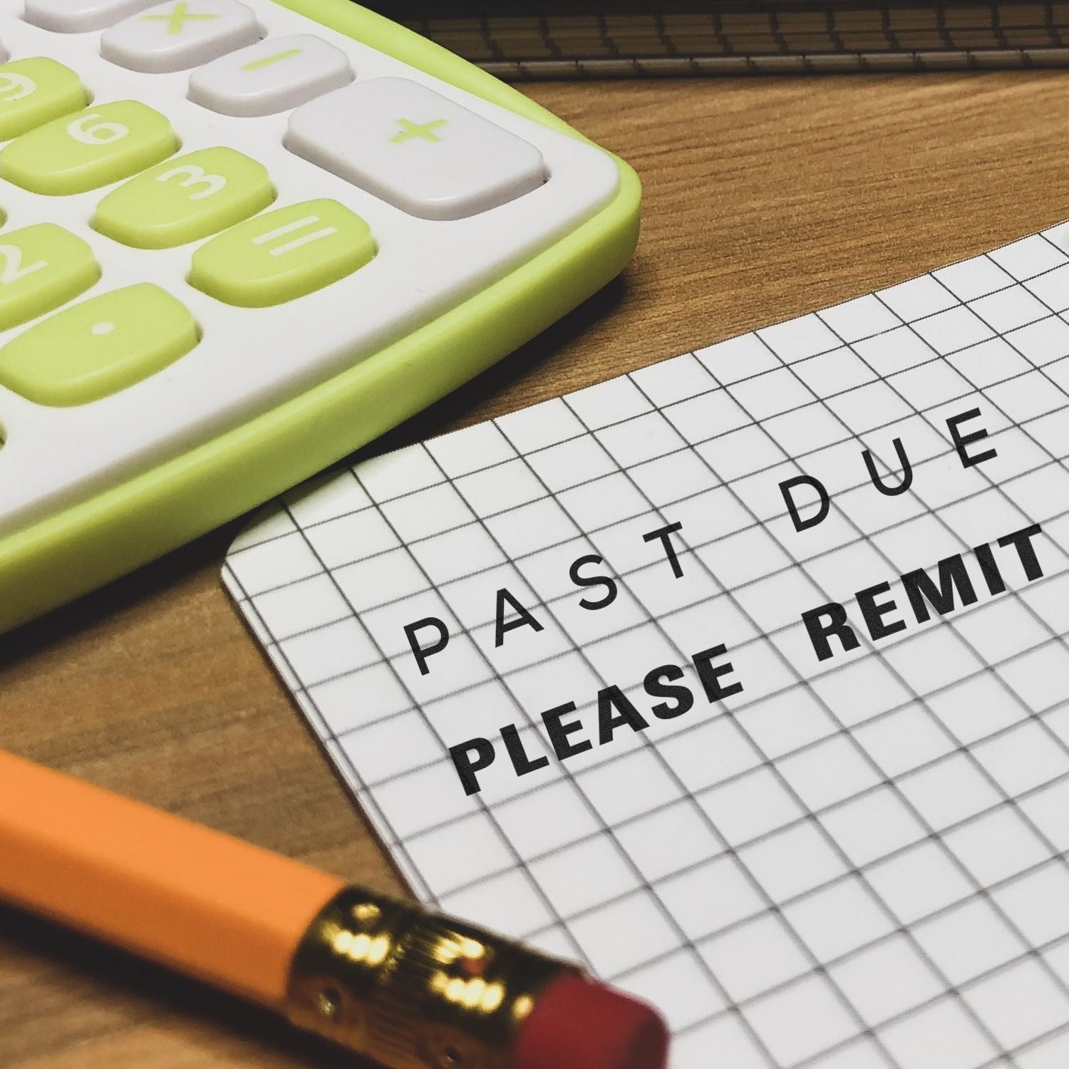 A Self Inking Past Due Please Remit Stamp is used on a grid paper next to a yellow calculator and a pencil on a wooden desk.