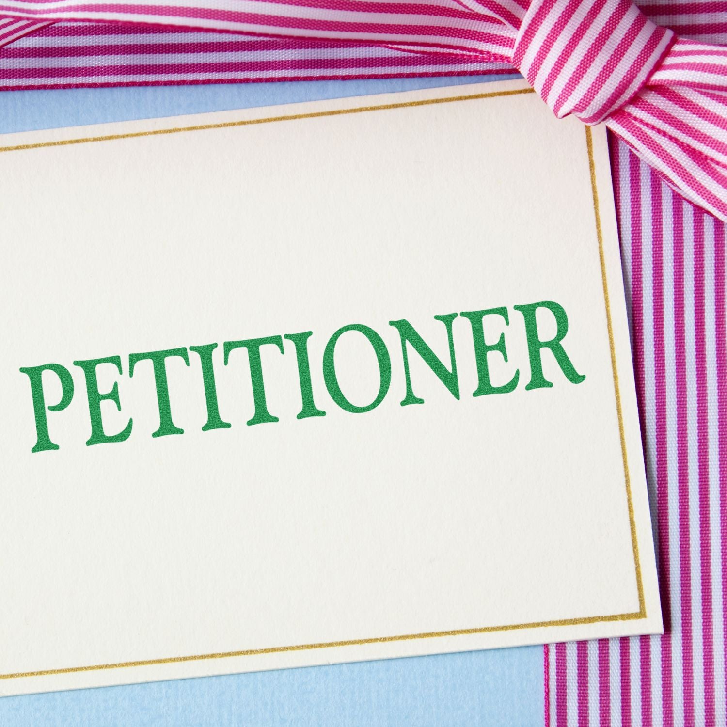 Large Petitioner Rubber Stamp in use, stamping PETITIONER in green ink on a white card with pink and white striped ribbon.