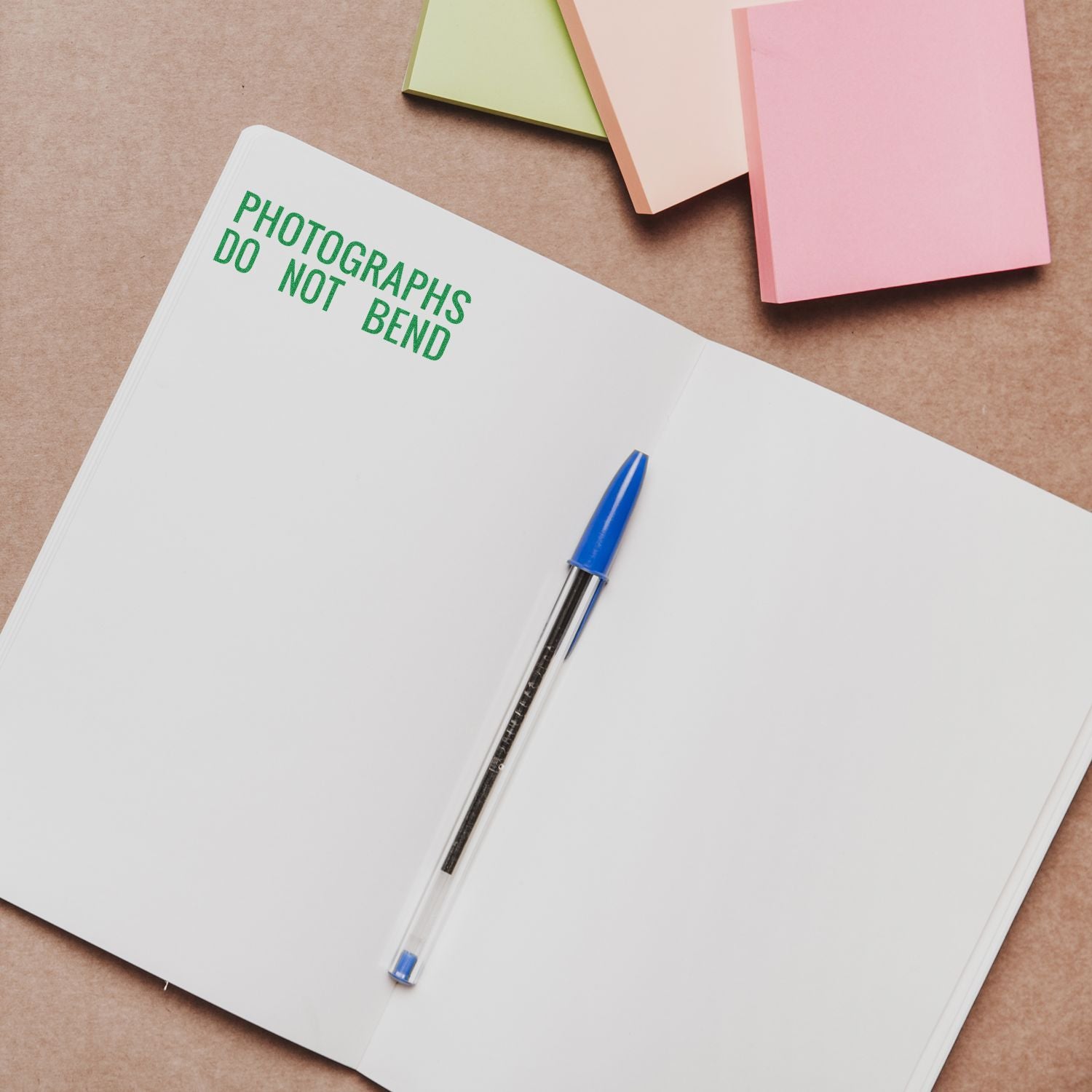 Open notebook with Photographs Do Not Bend stamped in green, a blue pen, and colorful sticky notes. Product: Large Photographs Do Not Bend Rubber Stamp.