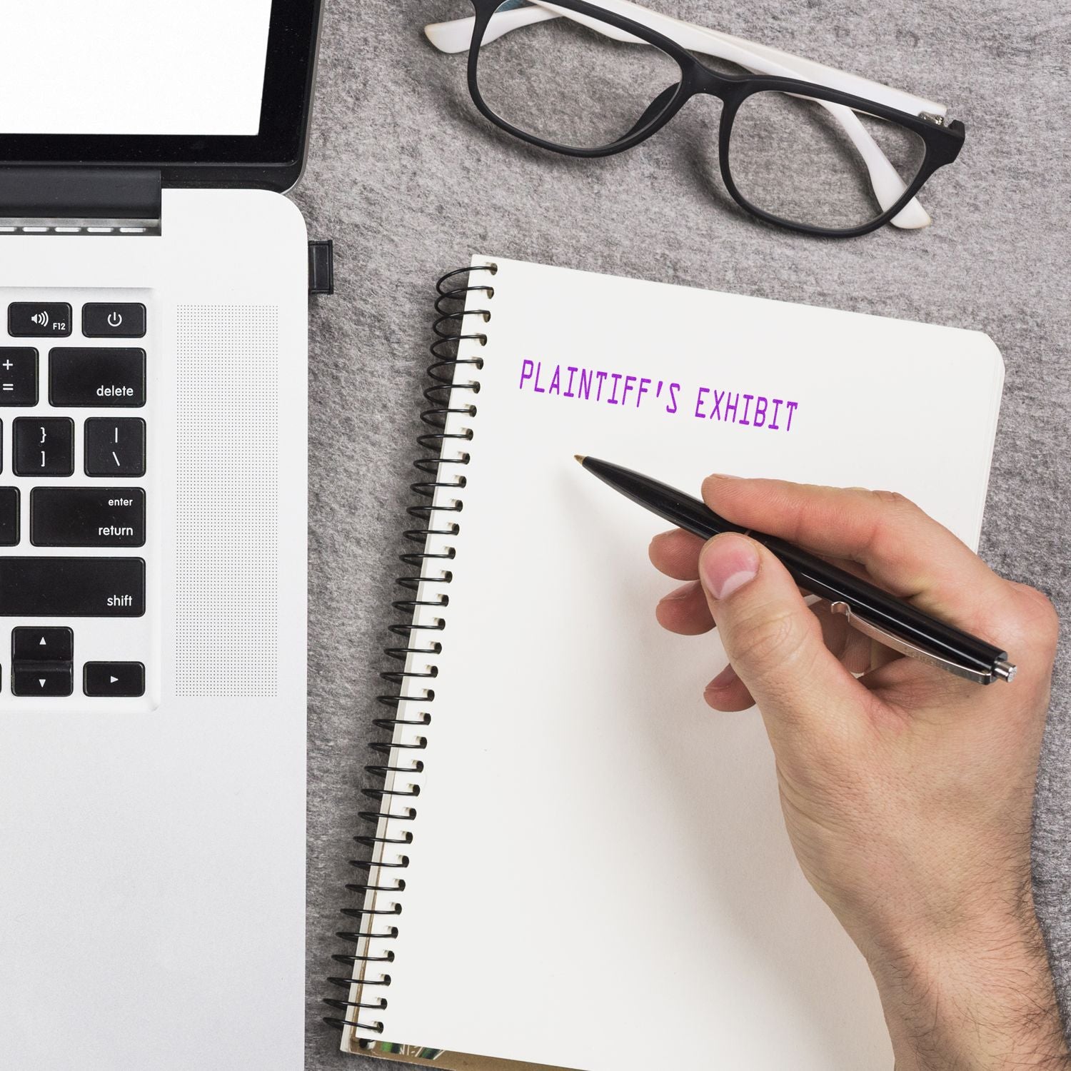 A hand writing Plaintiff's Exhibit in a notebook next to a laptop and glasses, using a Slim Pre-Inked Plaintiff's Exhibit Stamp.