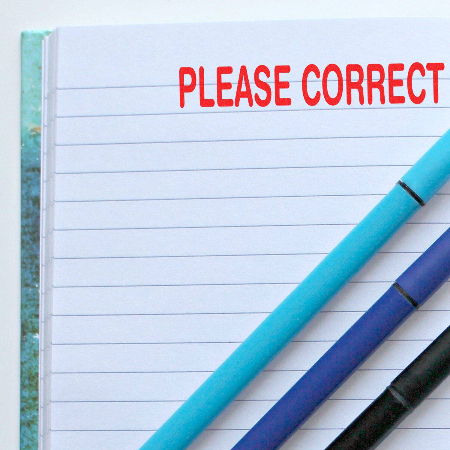 Large Pre-Inked Please Correct Stamp in red ink on a lined notebook page, with two pens placed diagonally across the page.