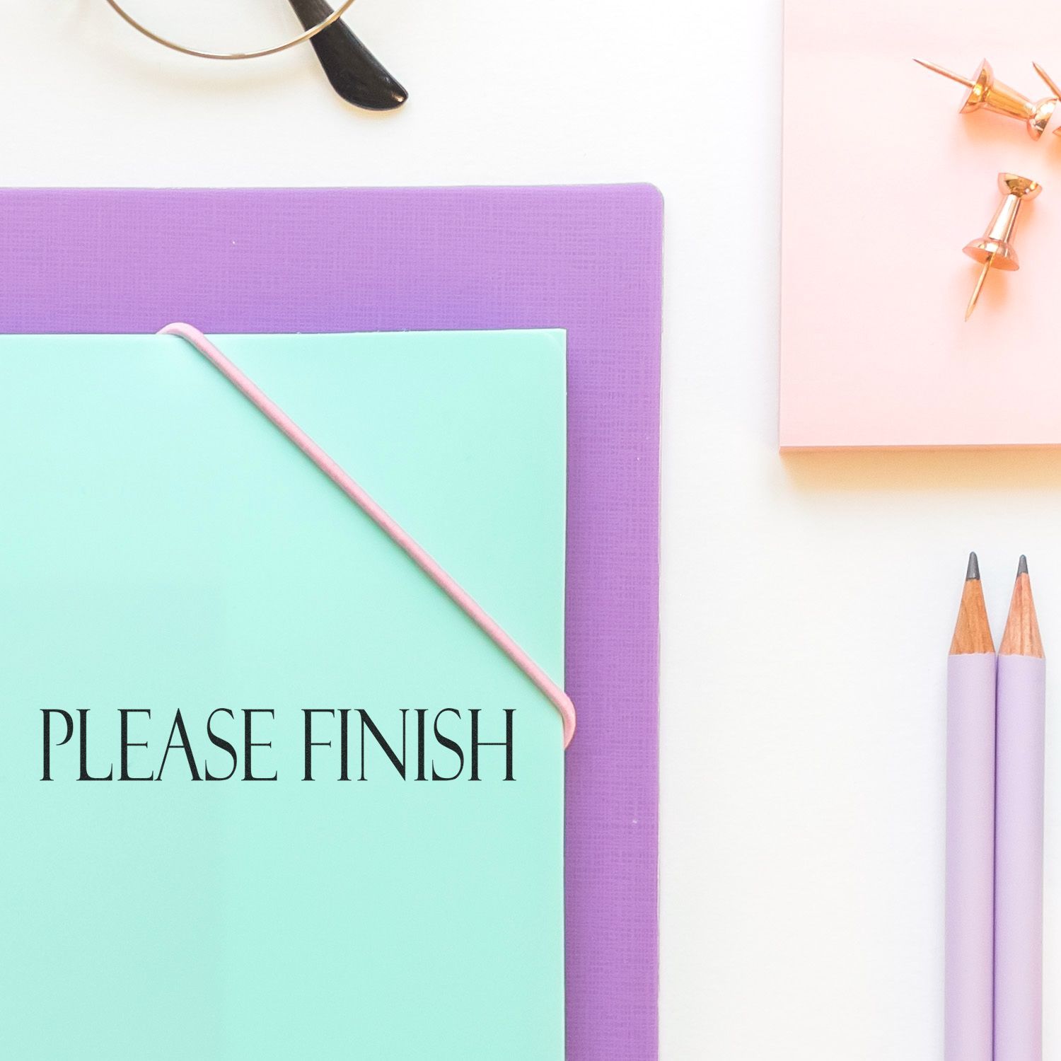 A Please Finish rubber stamp on a mint green folder, surrounded by a purple notebook, pencils, and push pins on a white desk.