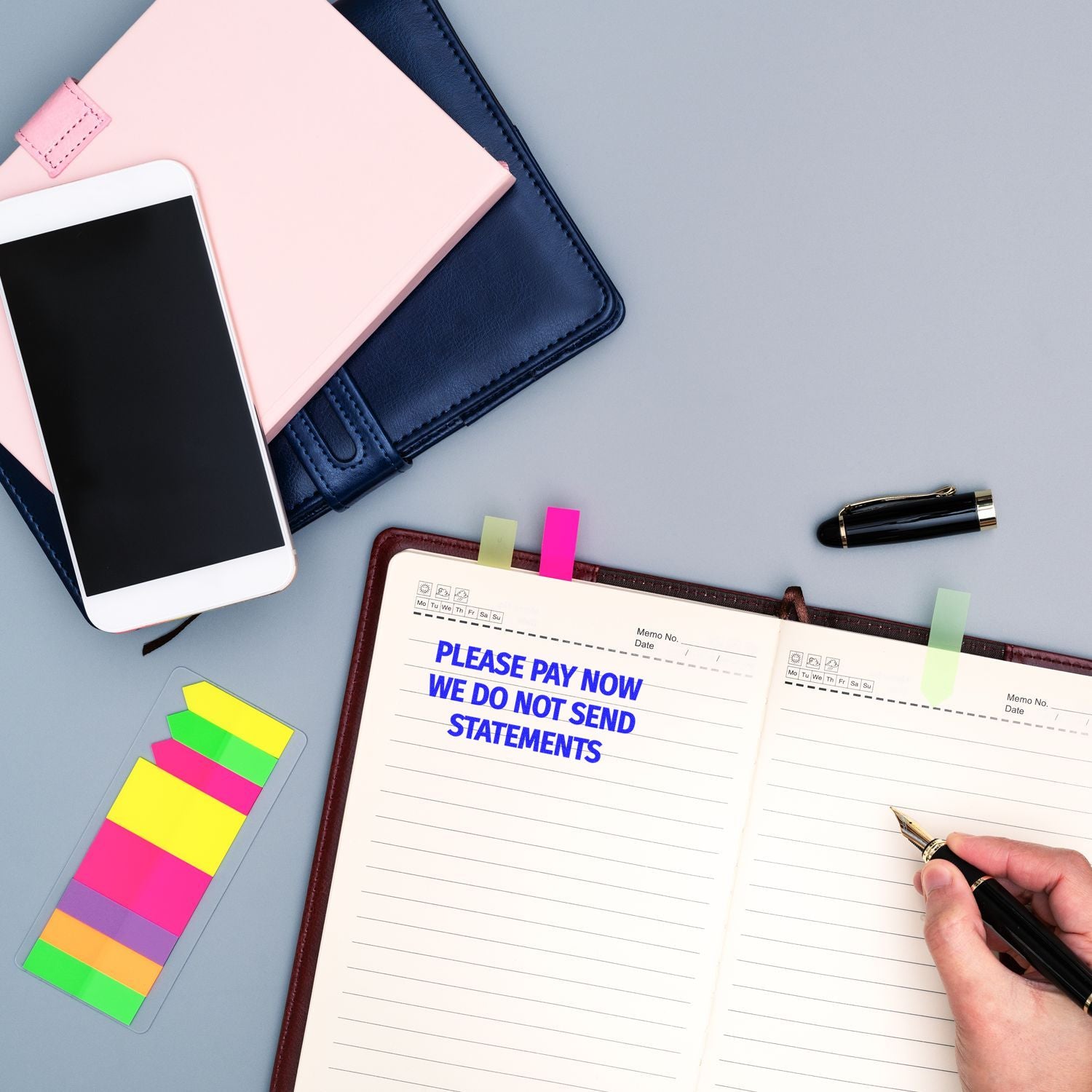 Person writing in a planner with a Large Please Pay Now No Statements will be Sent Rubber Stamp, surrounded by office supplies.