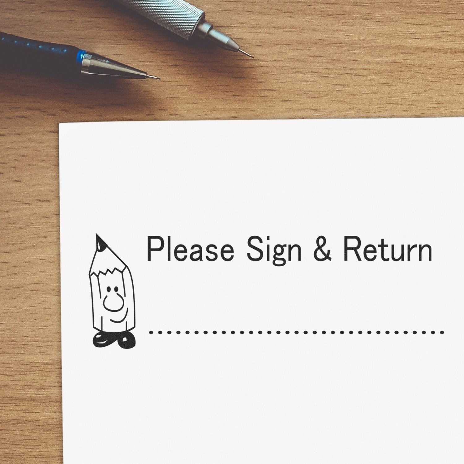 Large Self Inking Please Sign and Return Stamp on a piece of paper with a pencil illustration, placed on a wooden desk with two pens nearby.
