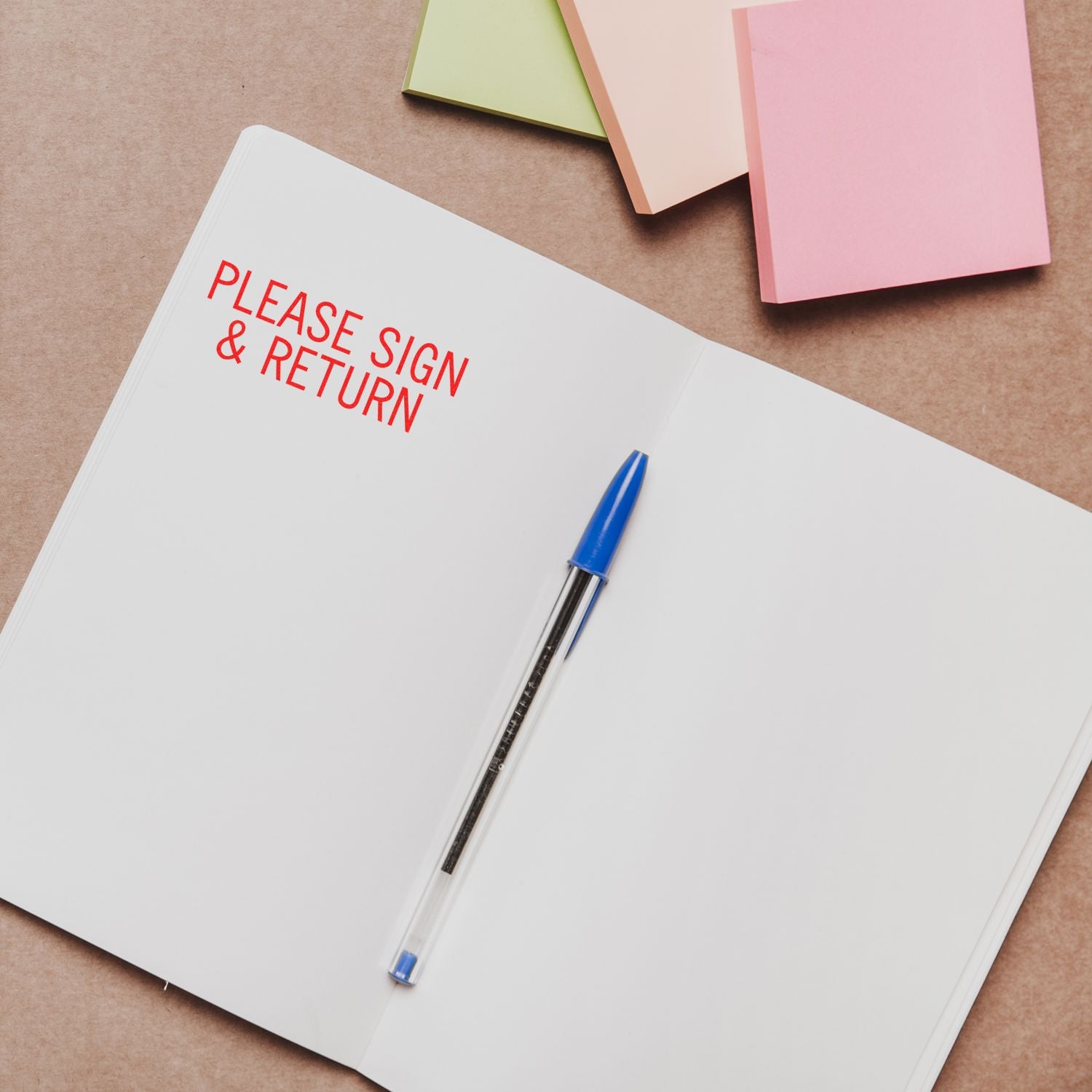 Open notebook with Please Sign & Return Rubber Stamp in red ink, blue pen placed on the pages, colorful sticky notes nearby.