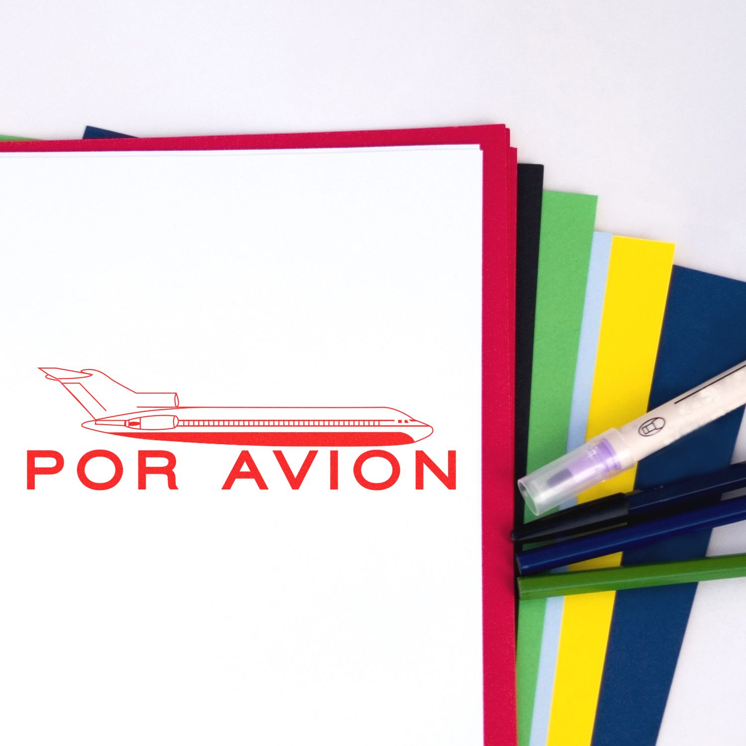 Self Inking Por Avion Stamp in red ink on white paper, with colorful sheets and pens in the background.