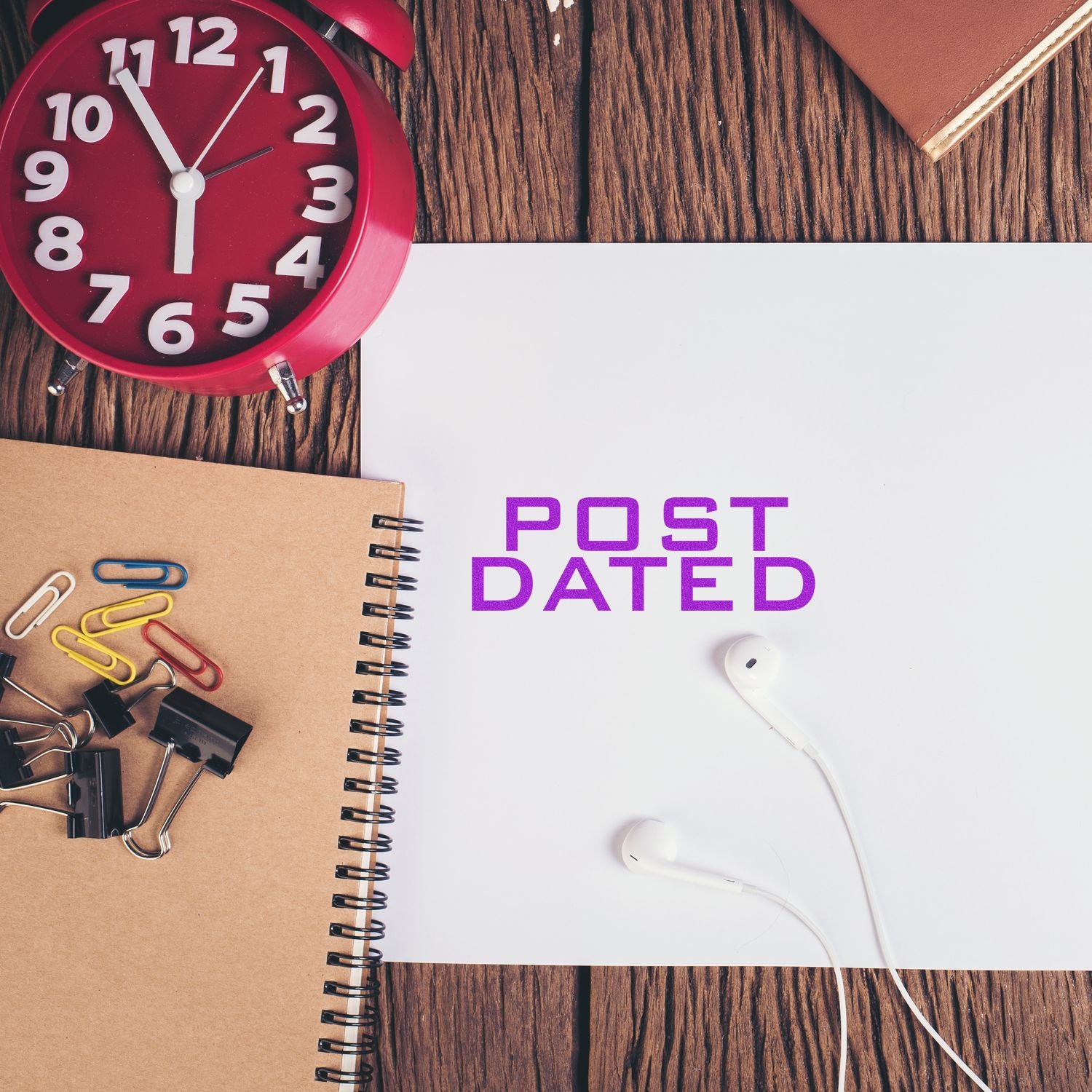 Post Dated rubber stamp on white paper with a red clock, notebook, paper clips, and earphones on a wooden desk.