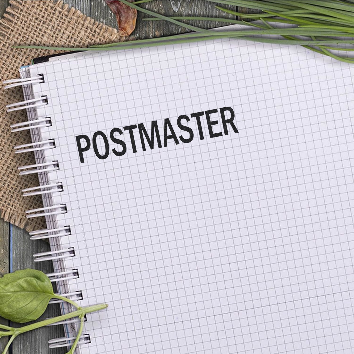 Notebook stamped with POSTMASTER using a Slim Pre-Inked Postmaster Stamp, placed on a wooden surface with leaves and burlap.