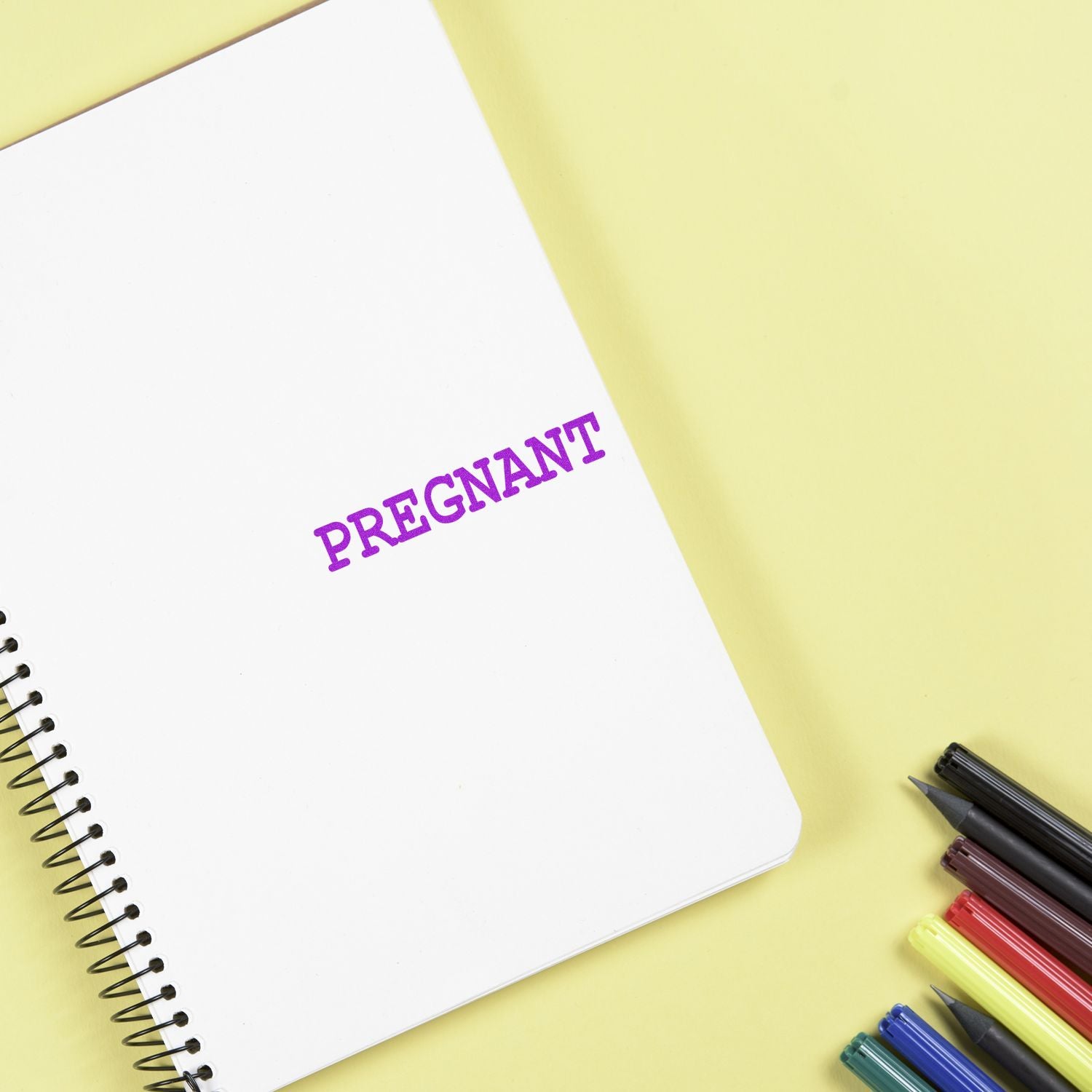 Notebook stamped with PREGNANT in purple ink using the Large Pre-Inked Pregnant Stamp, surrounded by colorful pens on a yellow background.