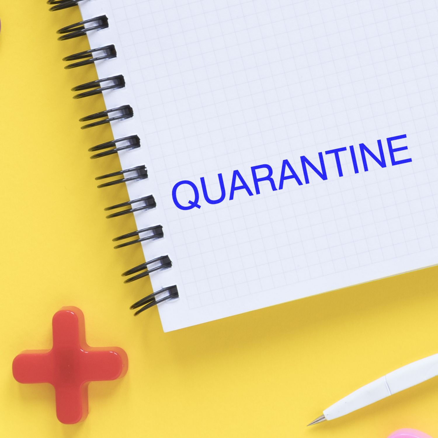 Quarantine rubber stamp impression on a spiral notebook with a red cross and a pen on a yellow background.