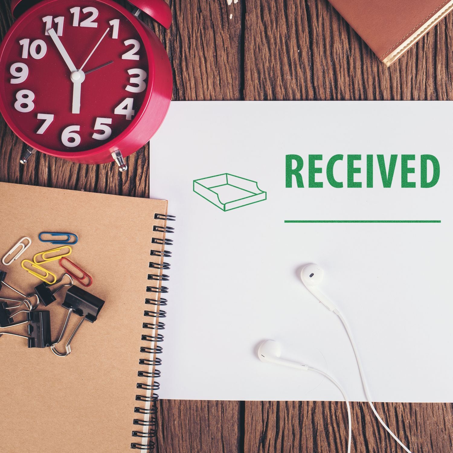 Desk with a red clock, notebook, paper clips, and earphones. Paper stamped with Large Received with Box Rubber Stamp in green.