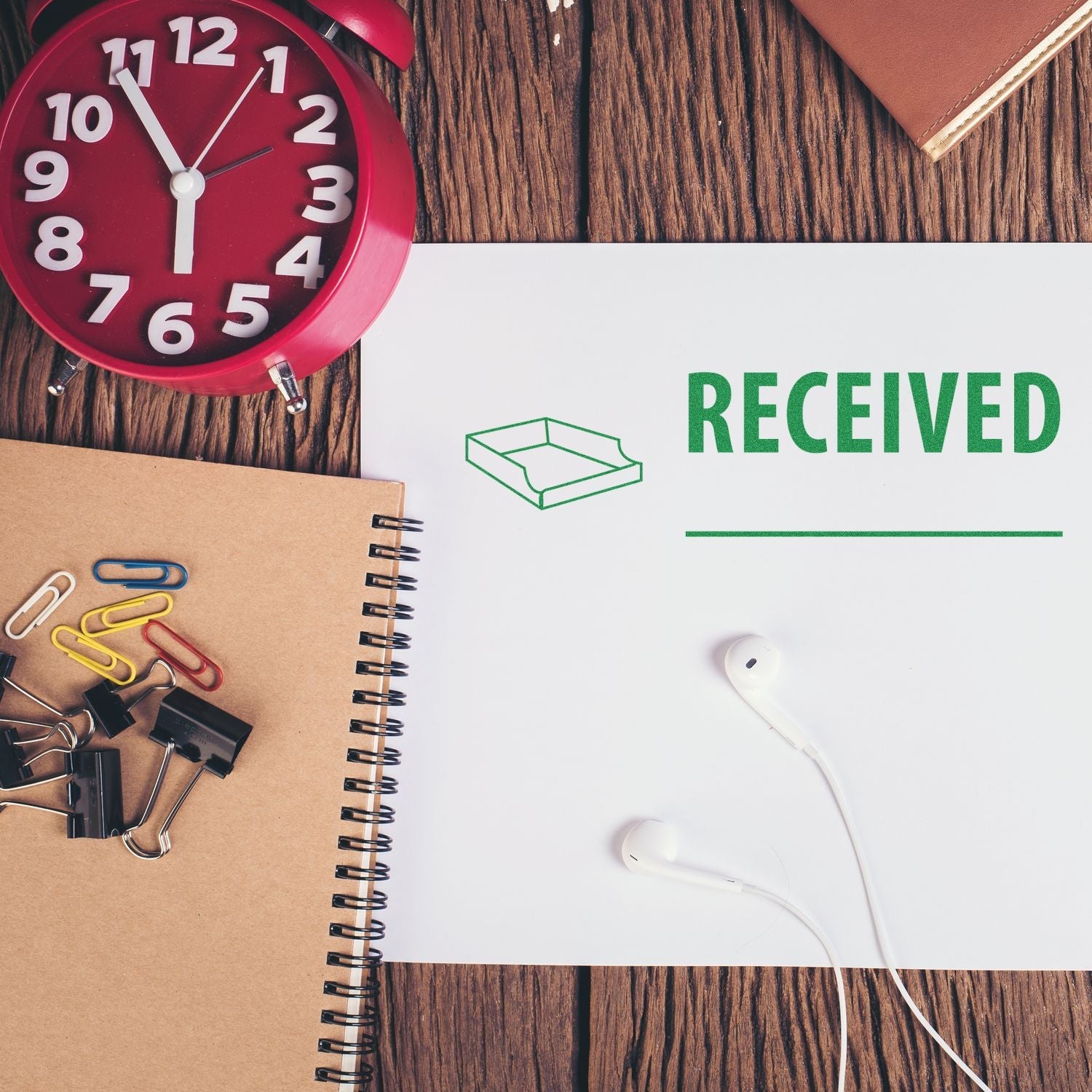 Self Inking Received with Box Stamp on paper, surrounded by a red clock, notebook, paper clips, binder clips, and earphones on a wooden surface.