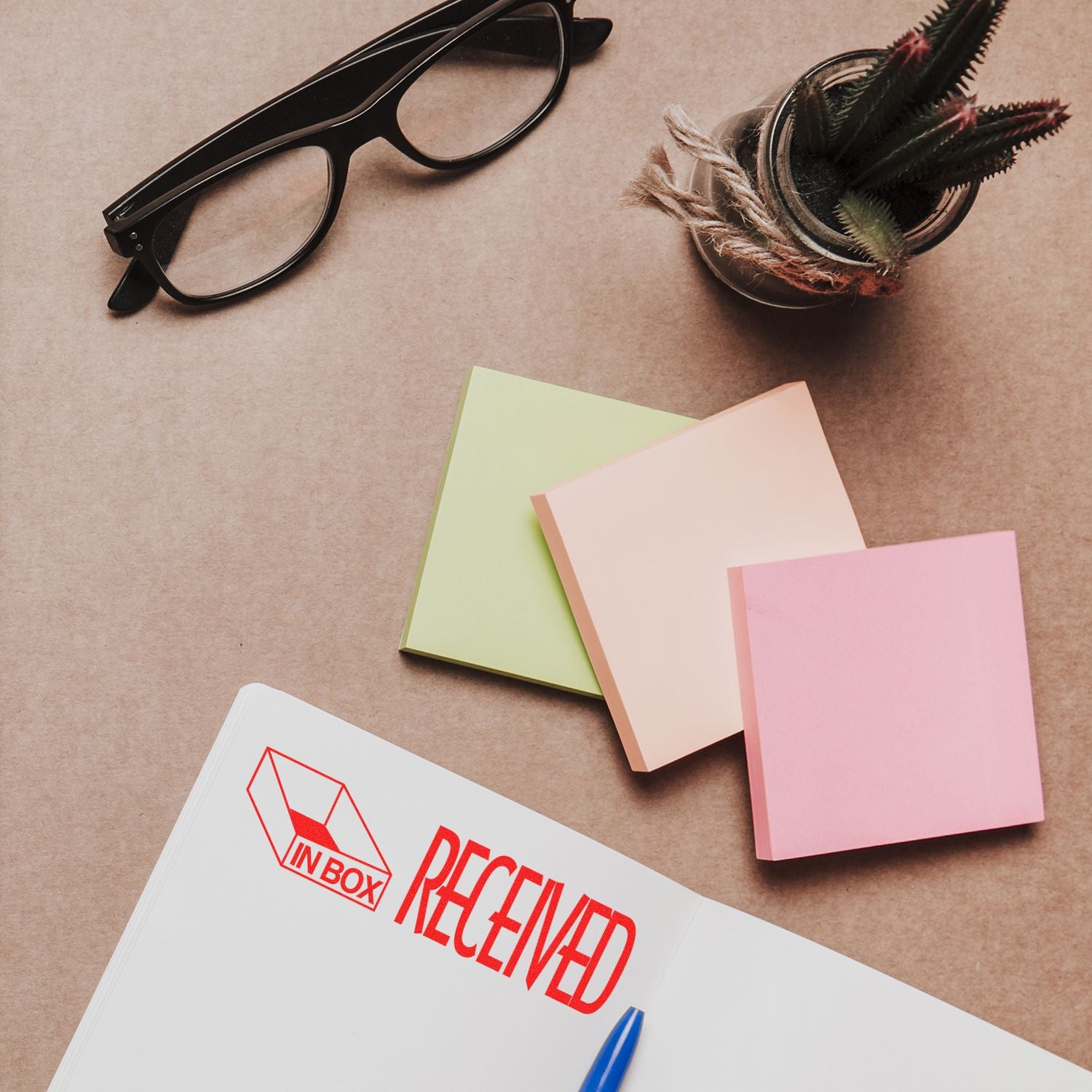 Received with Inbox Icon Rubber Stamp on a notebook, surrounded by sticky notes, a pen, glasses, and a small plant.