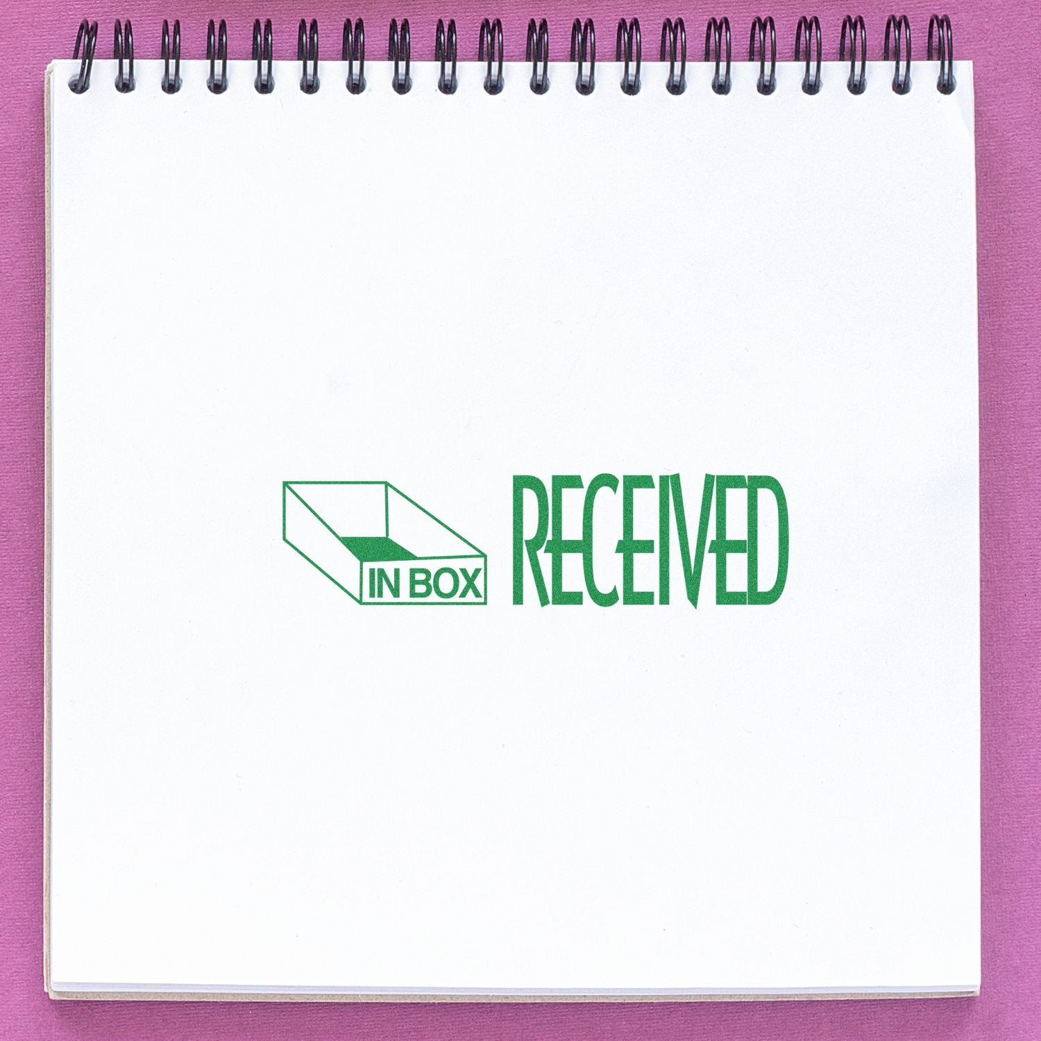 Slim Pre-Inked Received with Inbox Icon Stamp in green ink on a white spiral notebook, placed on a pink surface.